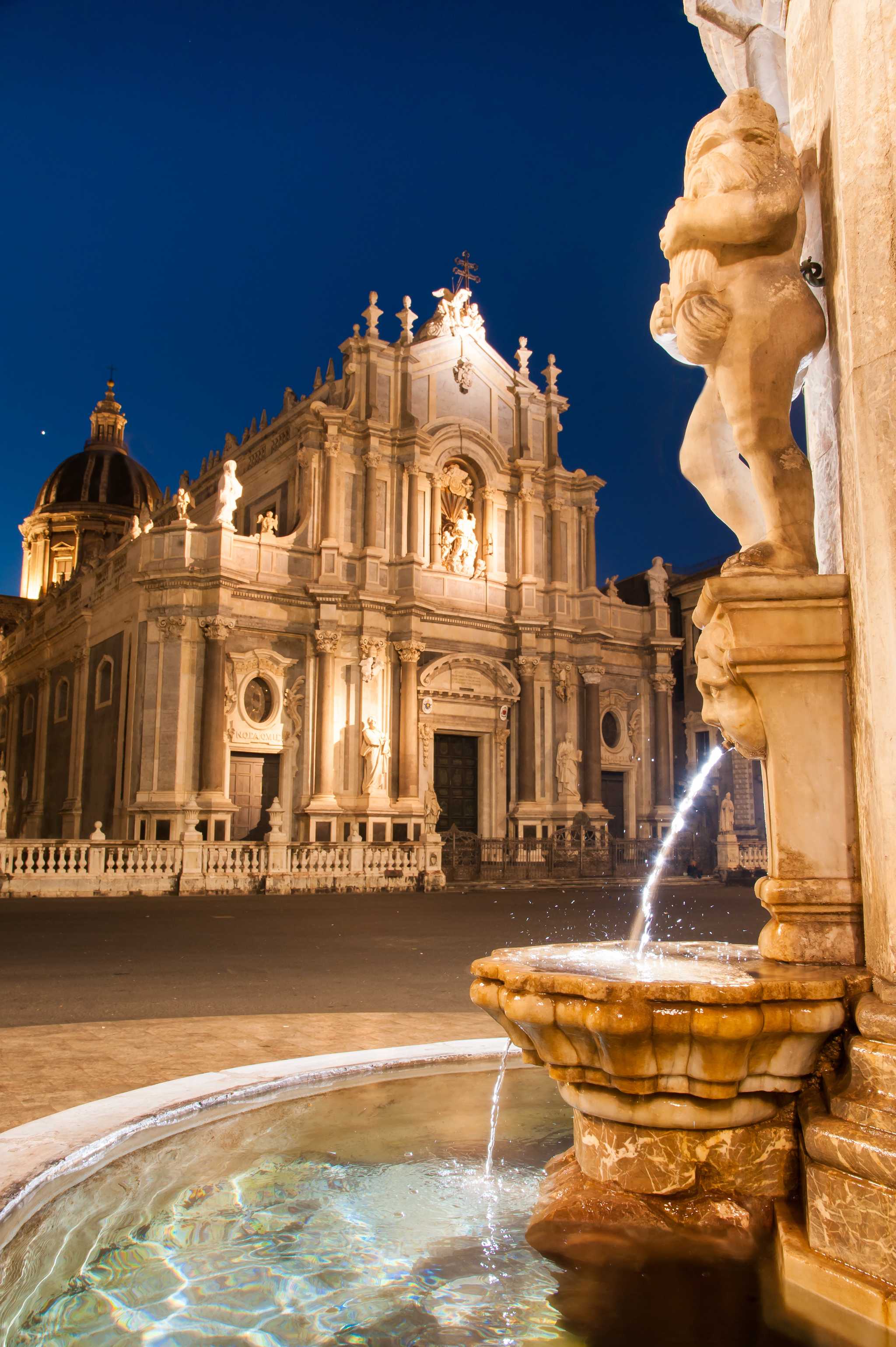 Cataniaâs stone Elephant Fountain illuminated at night, with the Cathedral of St. Agatha in the background.