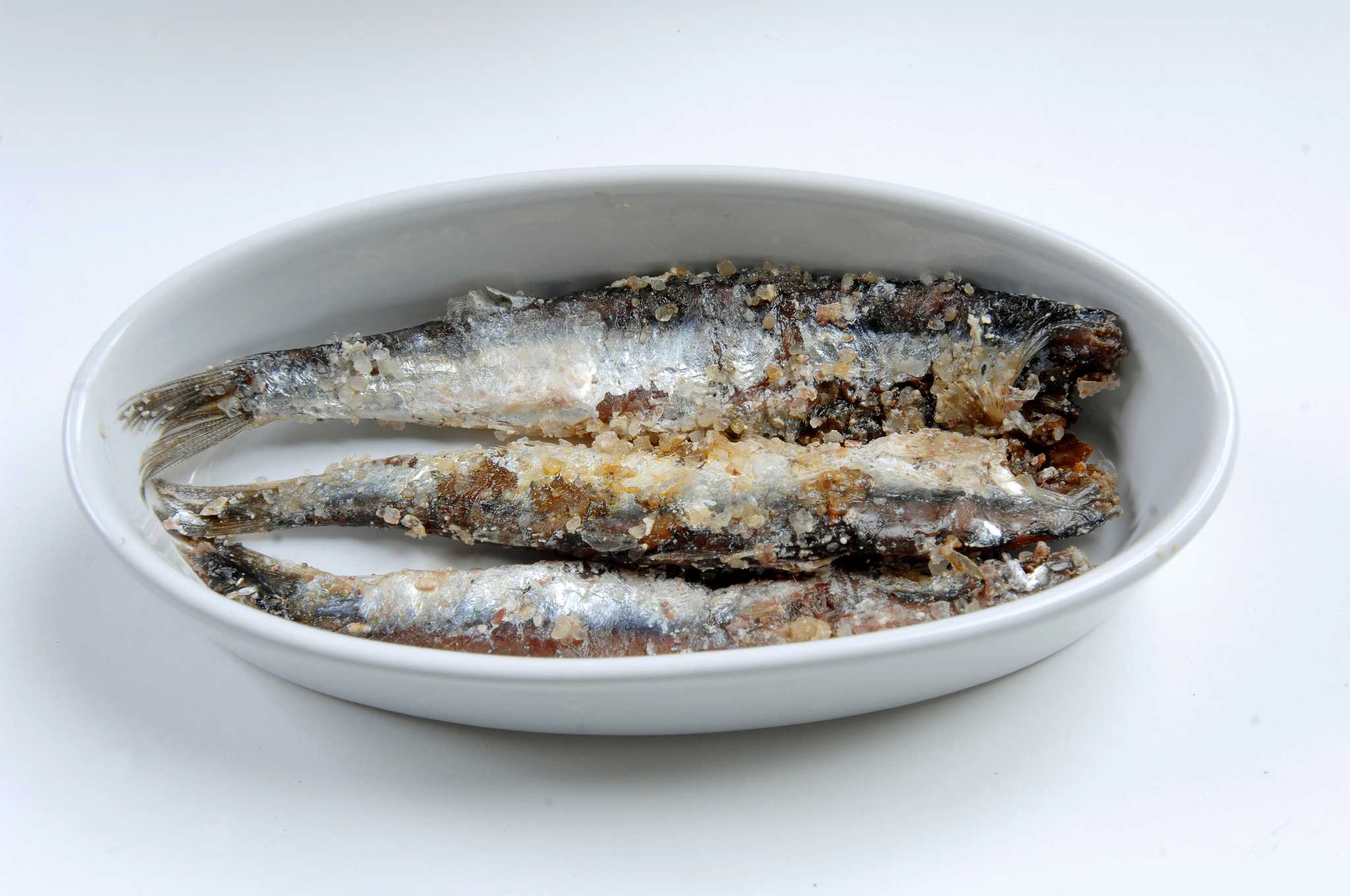 Whole salt-cured anchovies with silvery skin in coarse salt, in a small white dish.