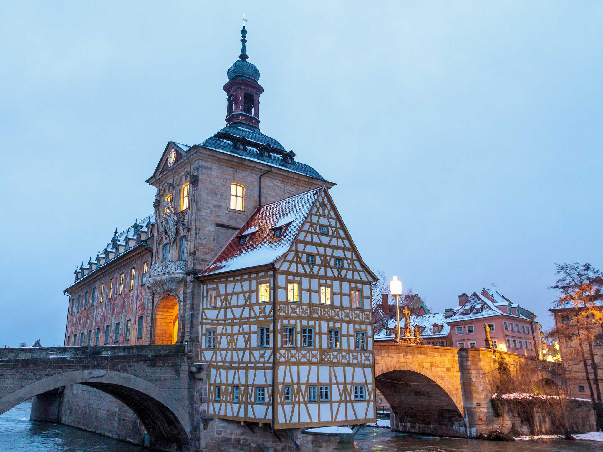 The Old Town Hall in Bamberg, a timber-framed section built on a stone bridge over a river during a snowy winter evening.