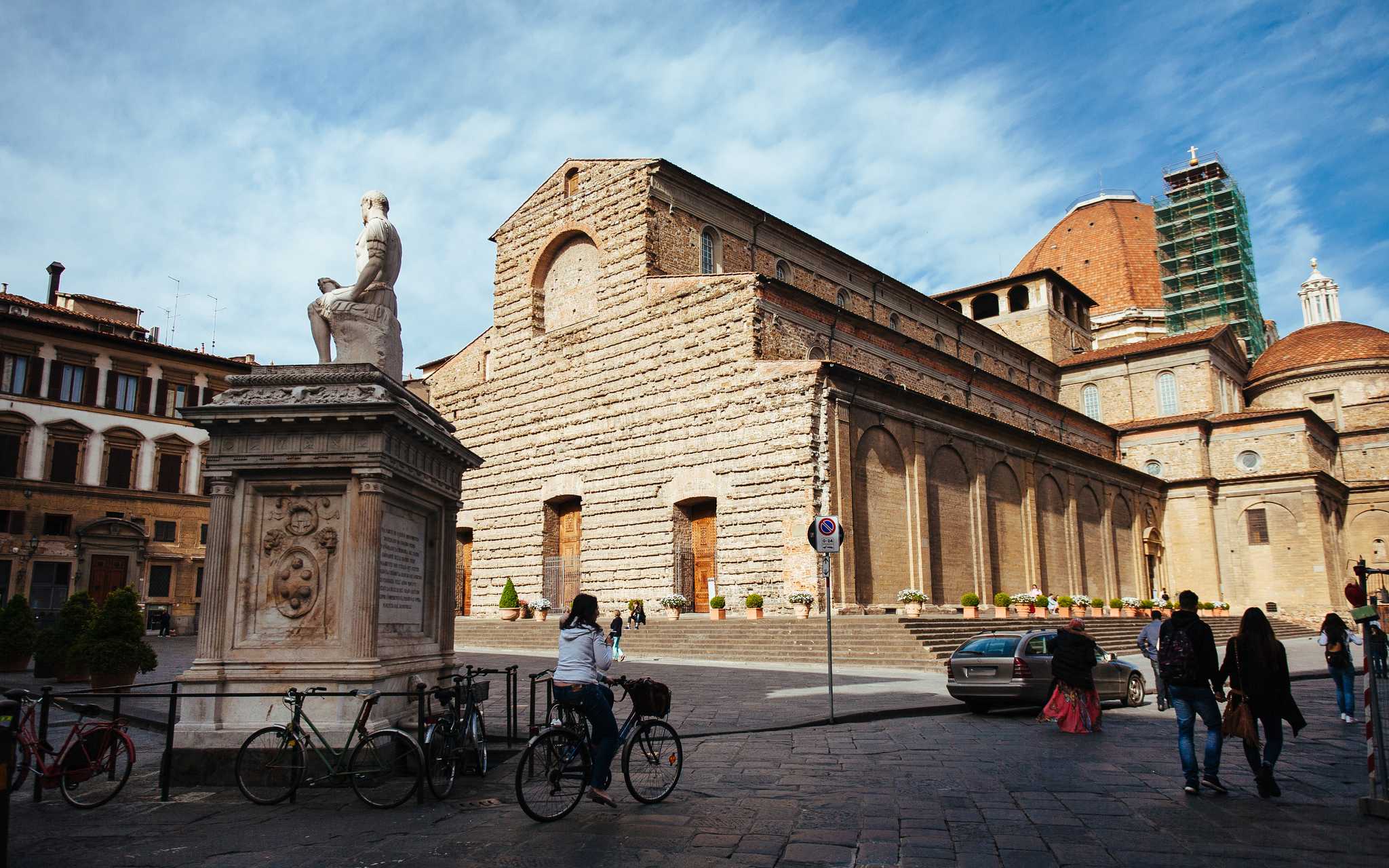 People walk and ride bicycles near the Basilica of San Lorenzo and a marble statue in a historic square under a blue sky.
