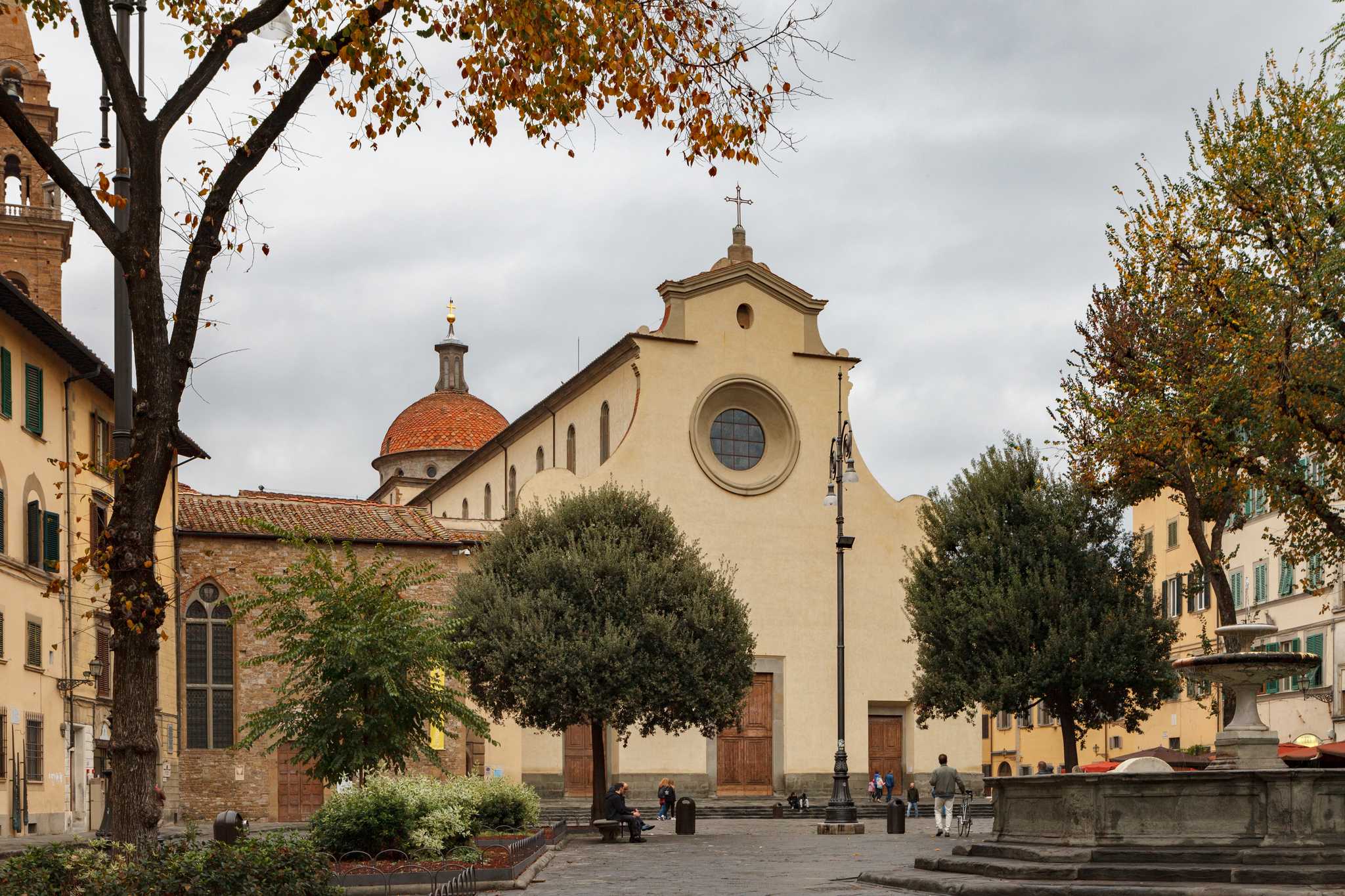 Basilica of Santo Spirito with a reddish dome in a town square, surrounded by trees, a stone fountain, and people walking on a plaza.