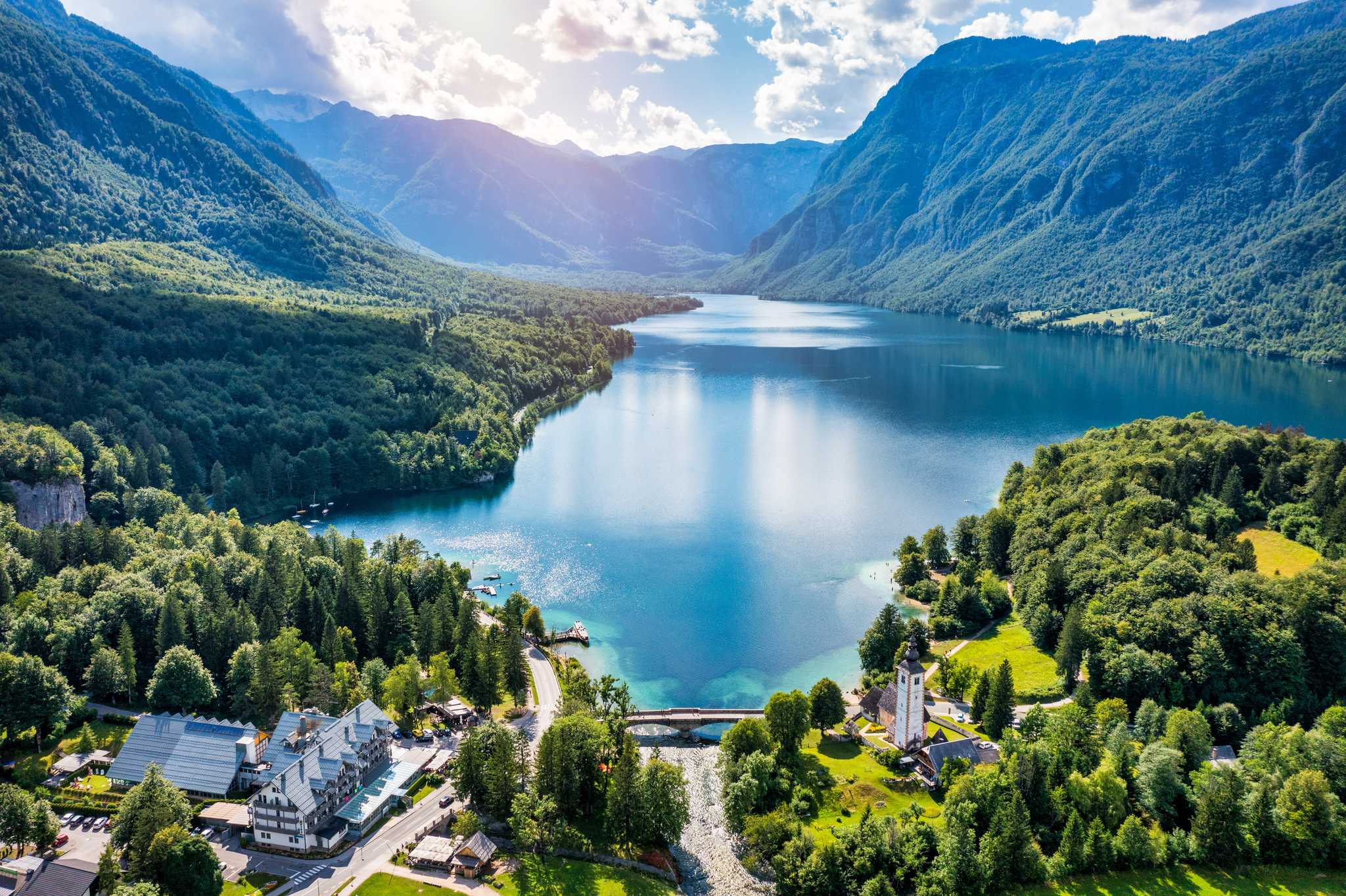 Lake Bohinj surrounded by lush green mountains, with a stone bridge, a church, and buildings along the shoreline.
