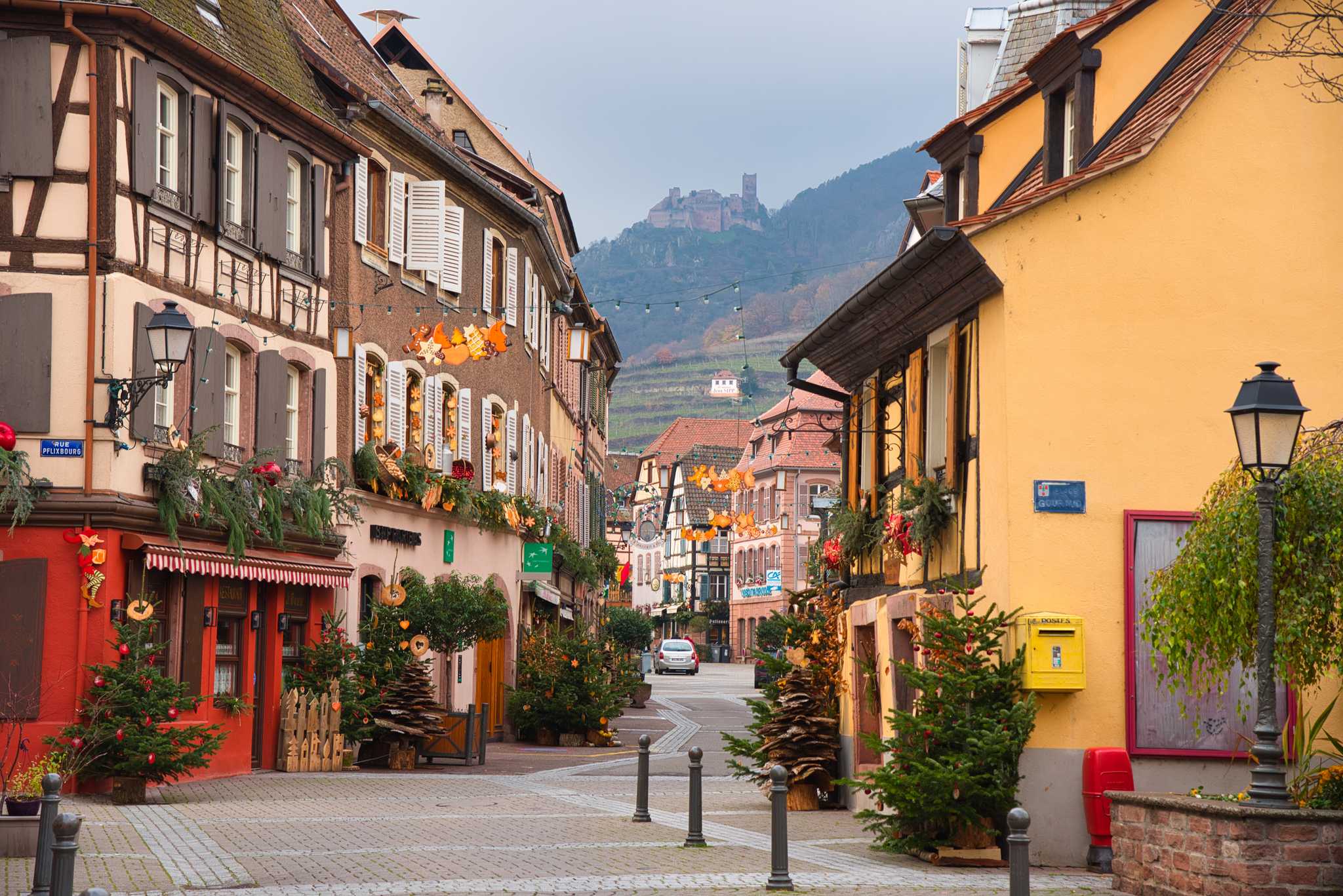 Alsatian village of Ribeauvillé with half-timbered buildings, Christmas decorations, small pine trees, and a distant castle on a hillside.