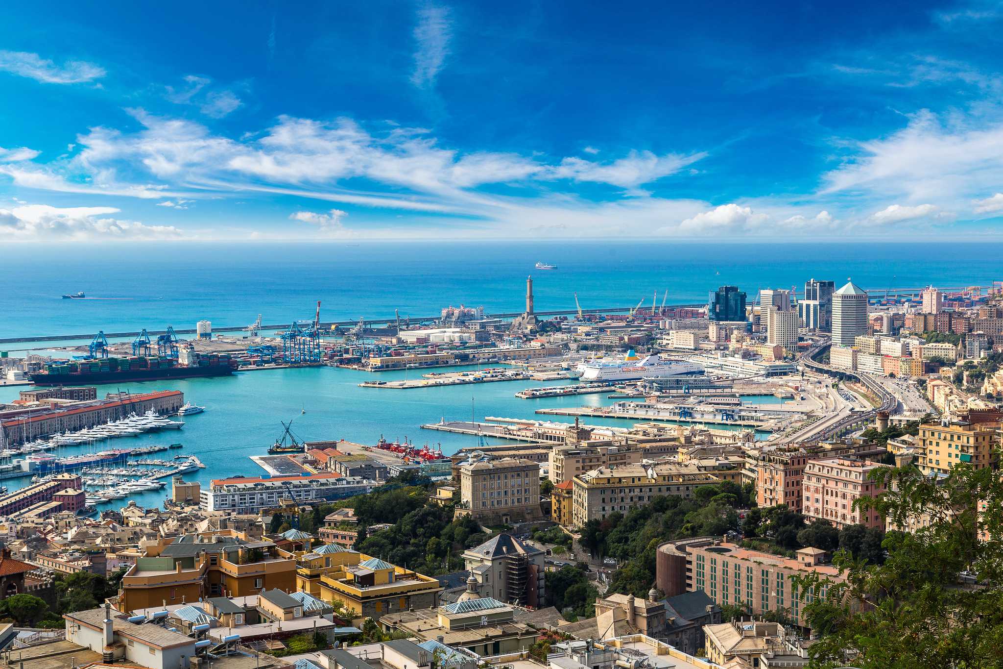 A high-angle view of the Genoa port, featuring a busy harbor with cargo ships, cruise liners, and city buildings under a blue sky.