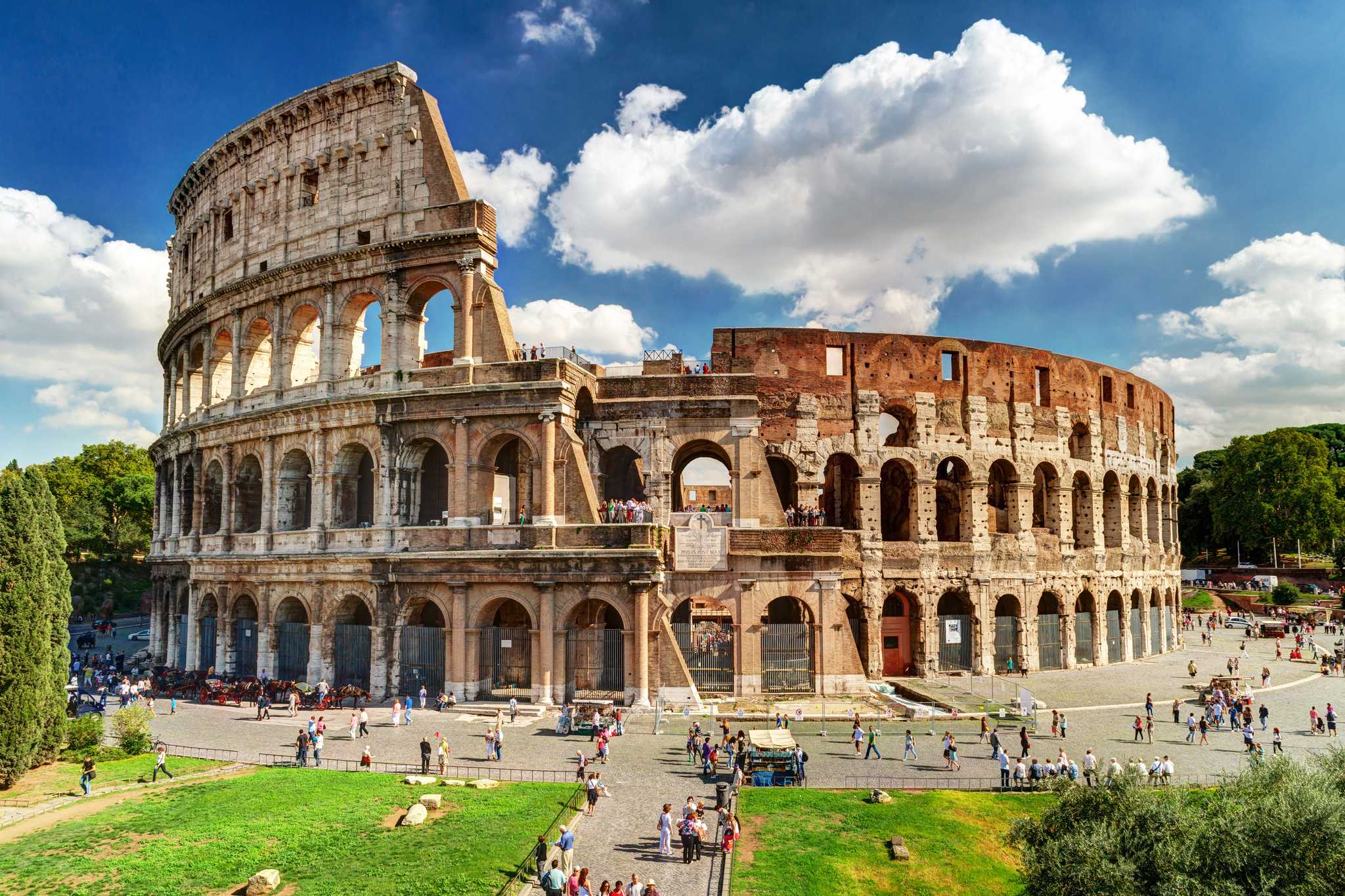 The Colosseum's arched tiers above a crowd-filled square under a bright, cloudy sky.