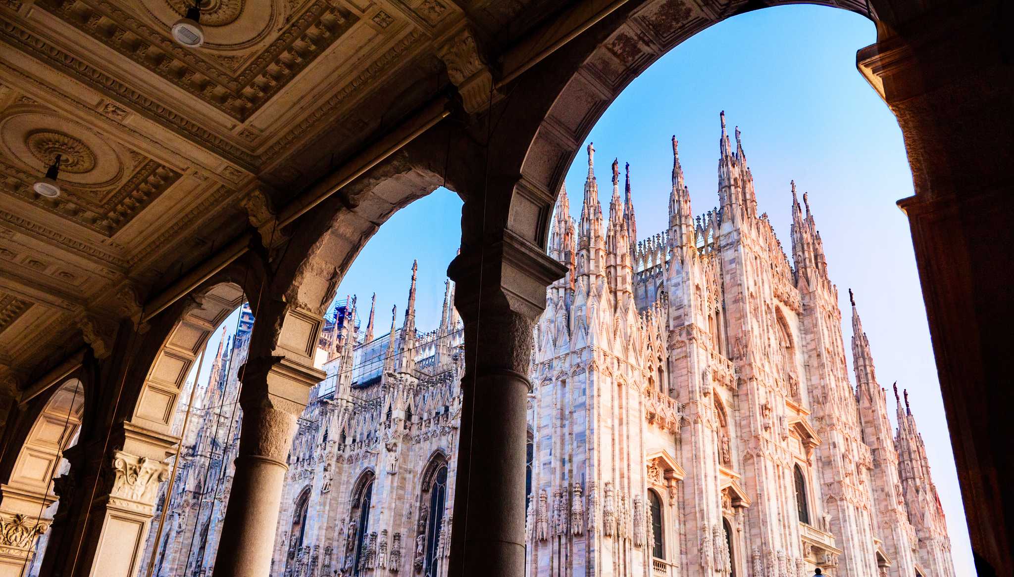 The white marble spires of Milan Cathedral, seen through the stone arches of a nearby arcade.