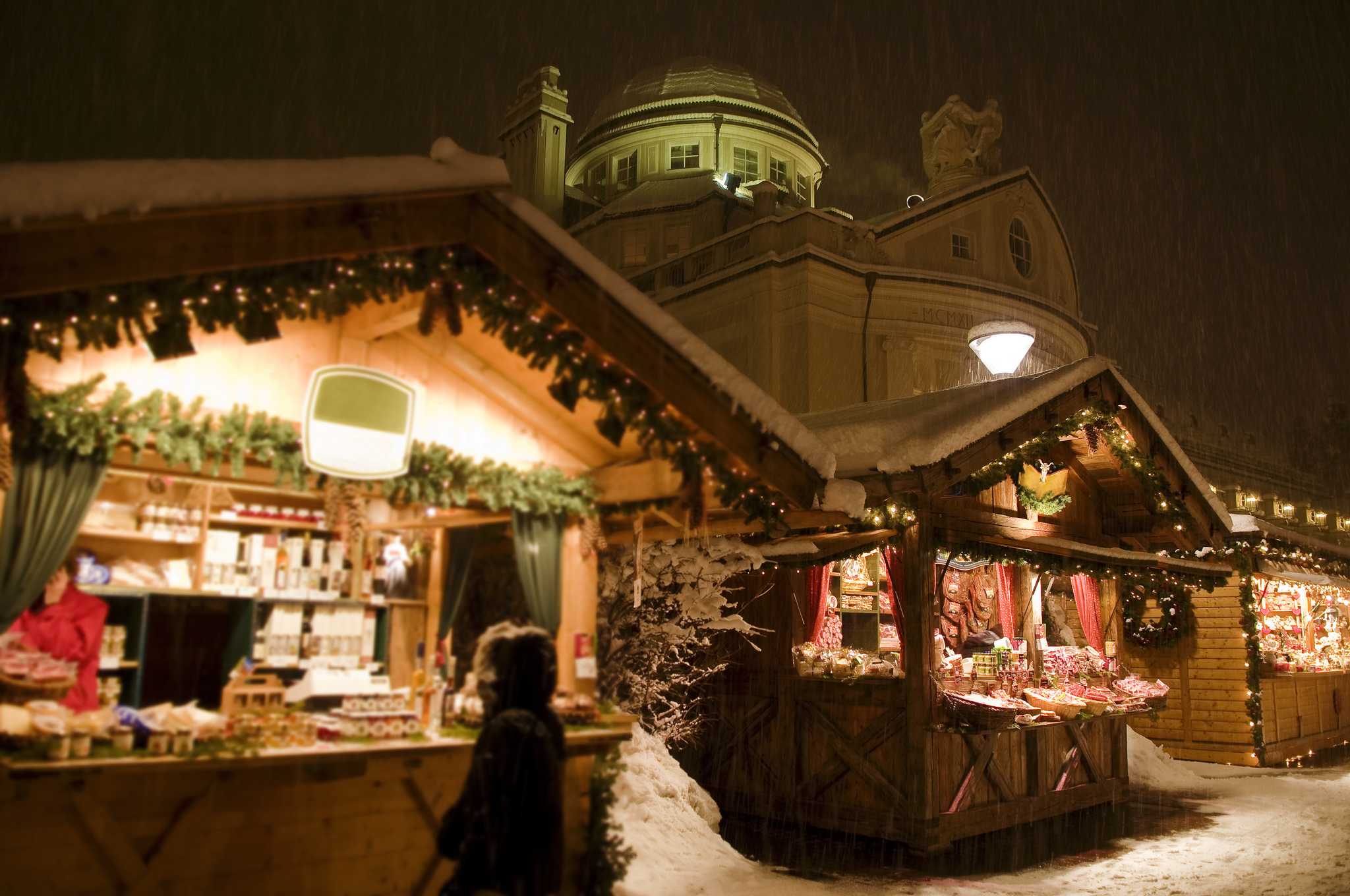 Lit wooden chalets at the Merano Christmas market during evening snowfall.