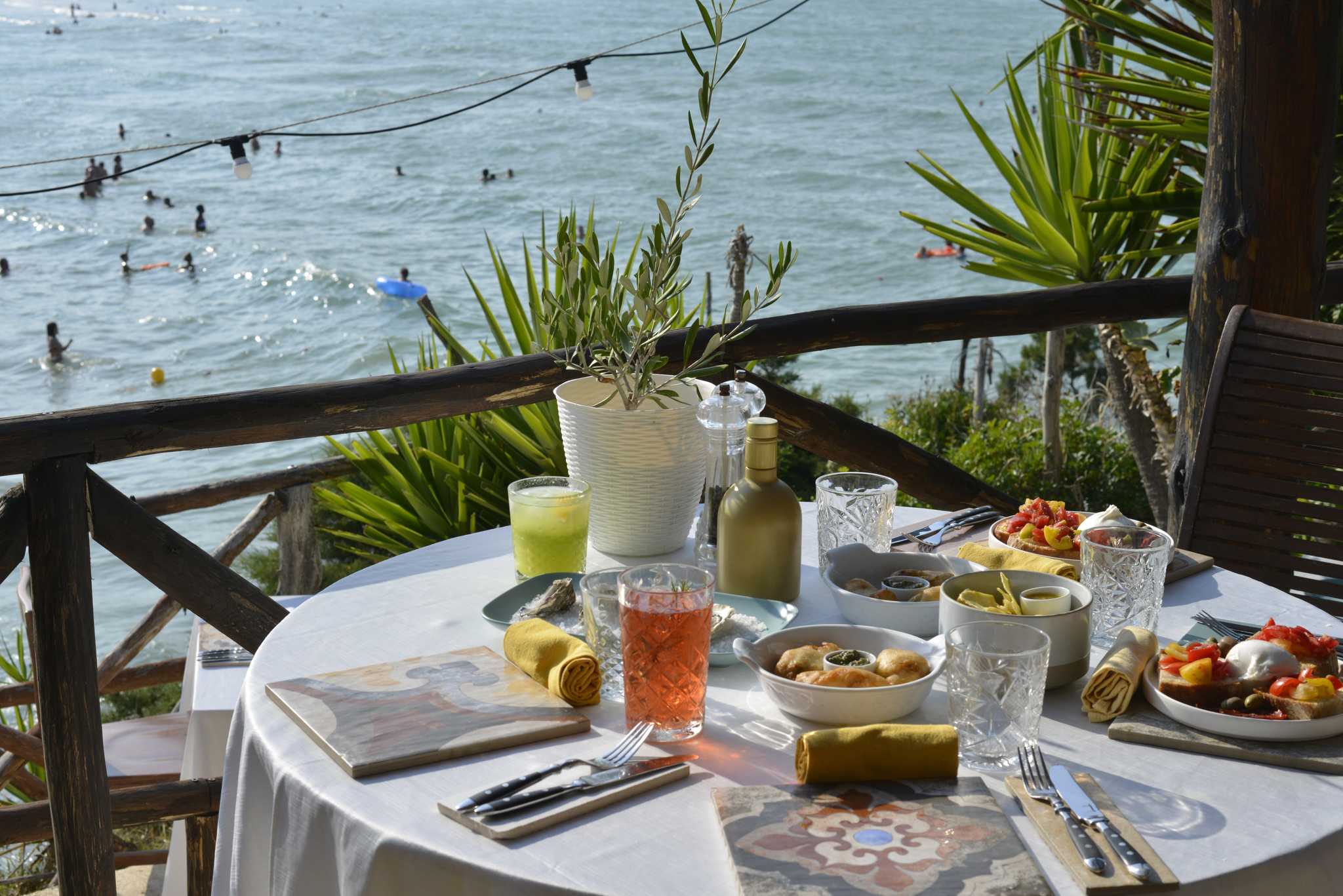 Seaside terrace table set with antipasti and drinks above swimmers in the water.