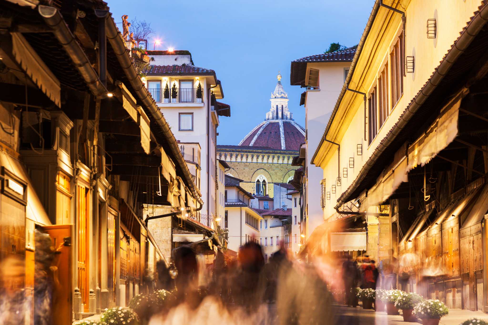 Crowds walk the Ponte Vecchio at dusk with the Florence Duomo beyond.