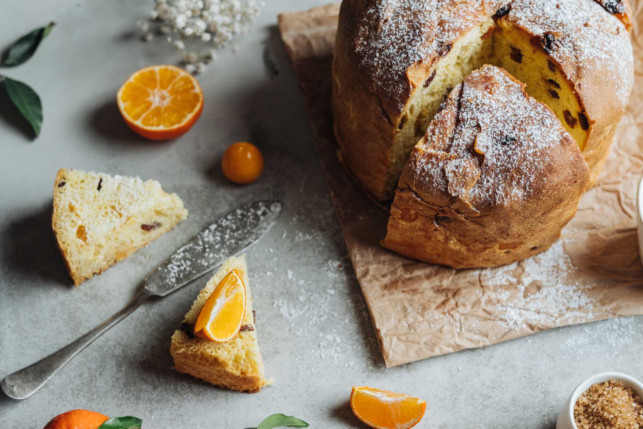 A tall domed sweet bread dusted with sugar and studded with candied fruit beside fresh oranges.