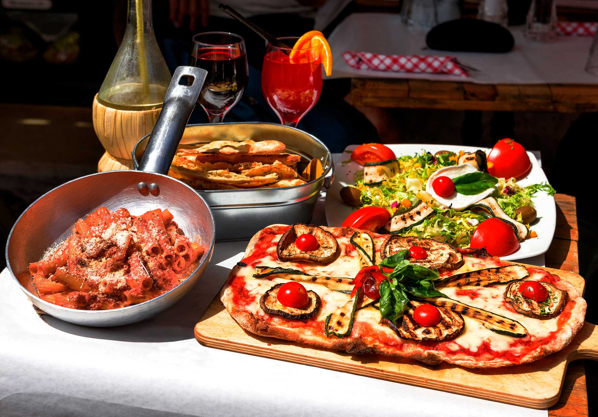 A sunlit trattoria table of pizza, pasta in a pan, salad, bread and drinks.