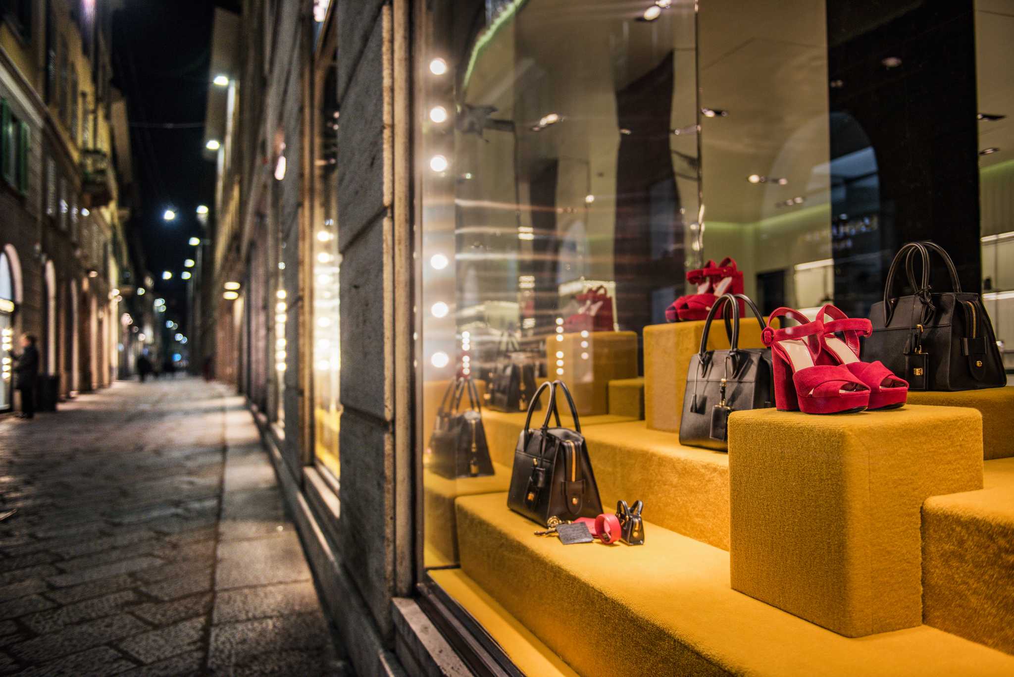 Designer handbags and heels lit in a boutique window on a shopping street at night.
