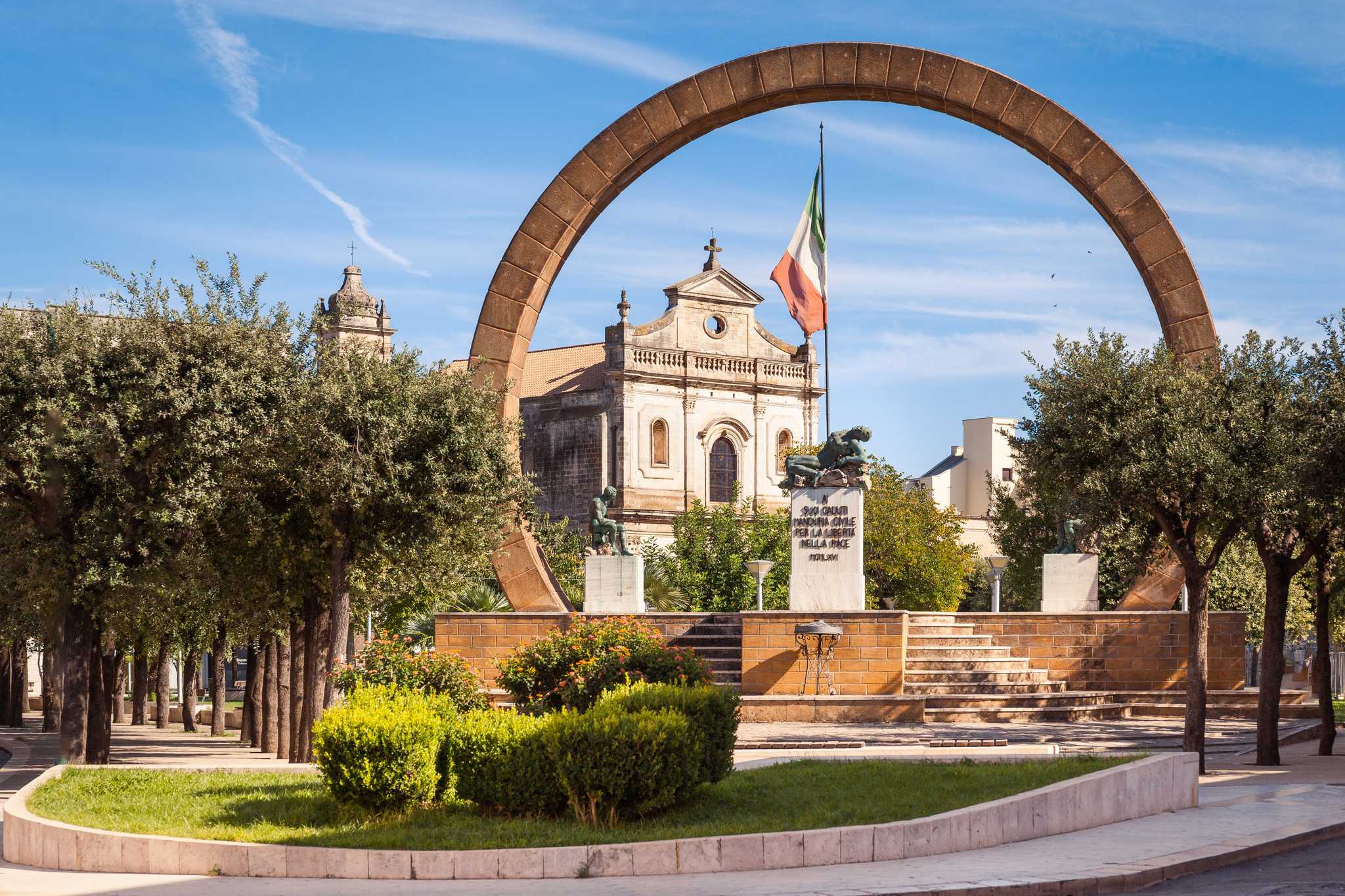 Stone arch framing a baroque church and war memorial in a tree-lined square.