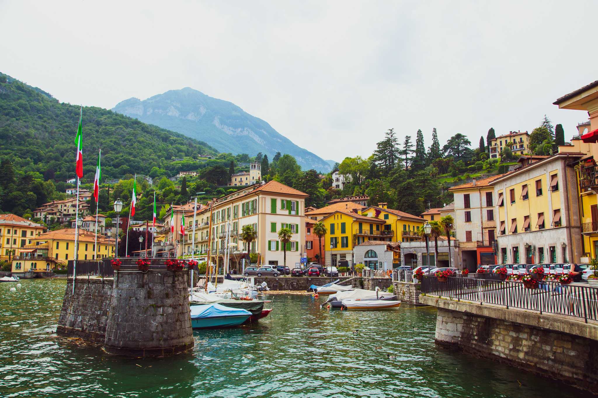 Boats moored in Menaggio's harbor on Lake Como below colorful houses and a mountain.