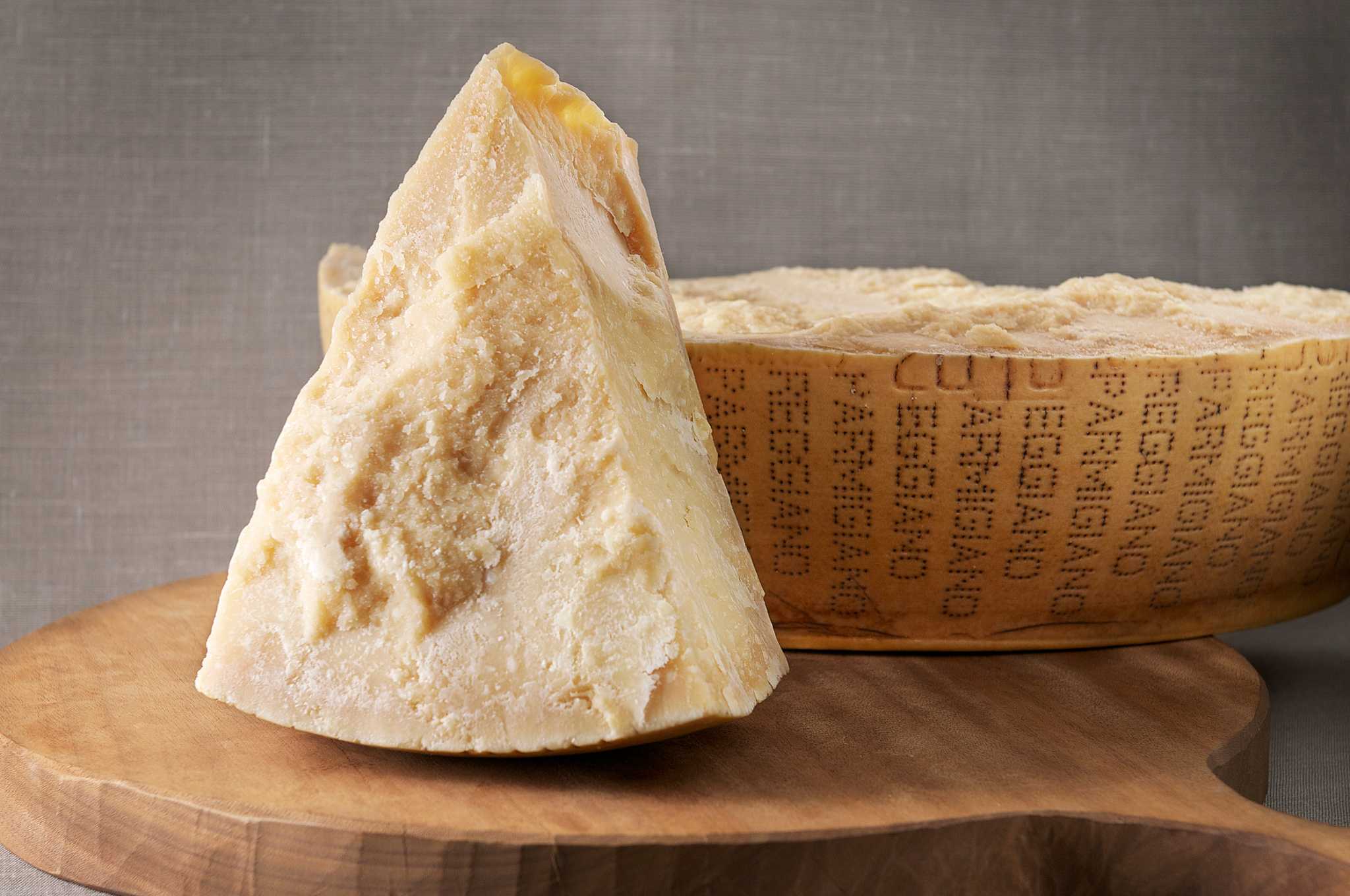 A slice of Parmigiano Reggiano cheese leaning against a wheel of cheese on a cutting board.