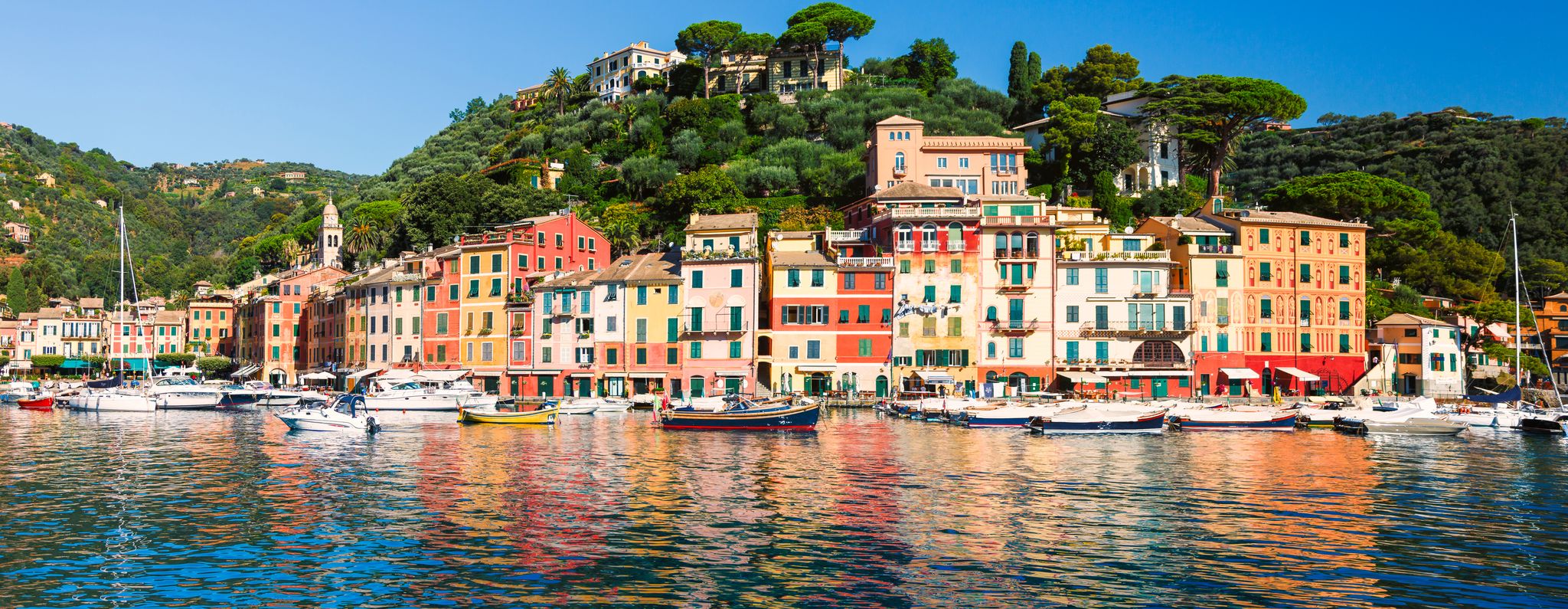 Colorful Portofino port with pastel buildings, boats, and green hills reflected in calm water under a bright blue sky.
