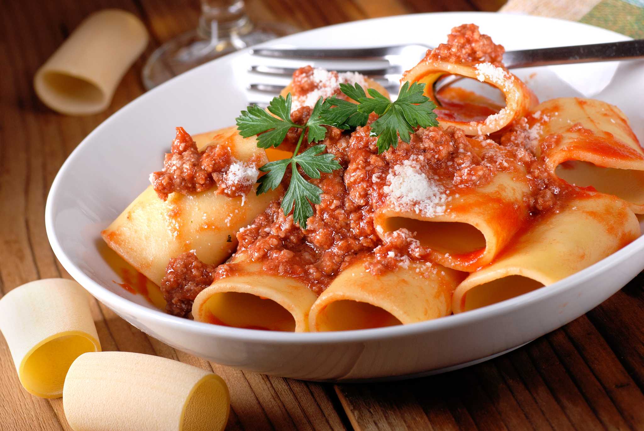 White bowl of wide tube pasta in rich meat ragu with grated cheese, dry pasta tubes alongside.