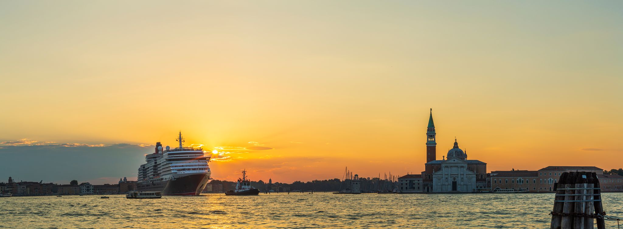 A large cruise ship on the water passing Venice, with a church dome and bell tower silhouetted against an orange sky.