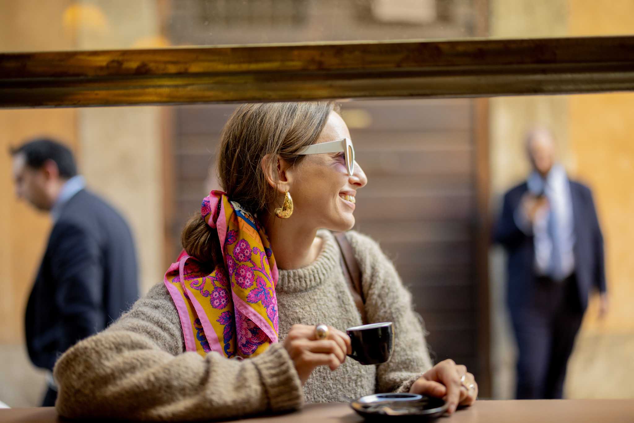 A woman visible through a window of a bar wearing a colorful headscarf and beige sweater holds a small espresso cup.