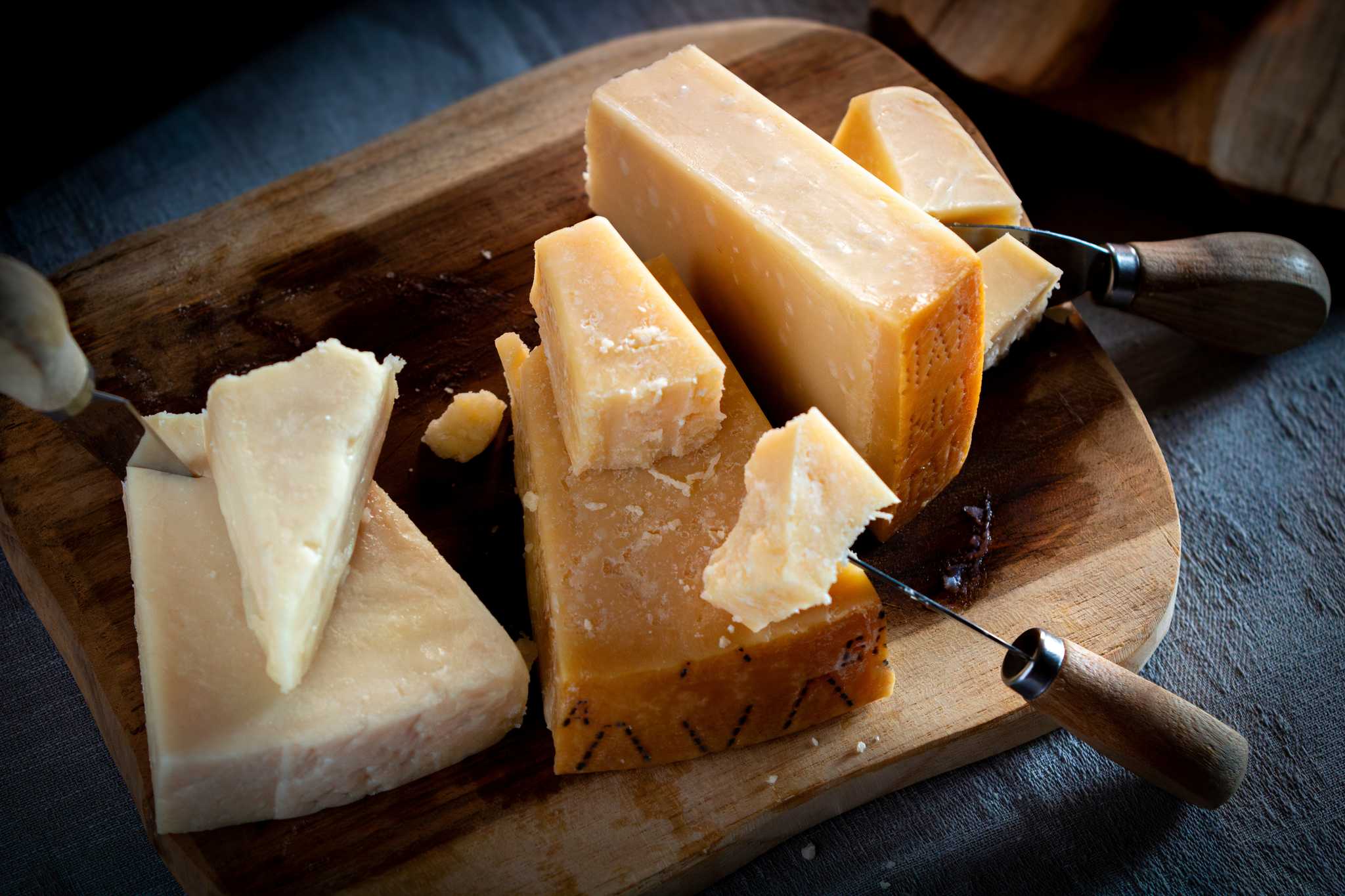 Three wedges of hard Italian cheese on a wooden board with cheese knives.