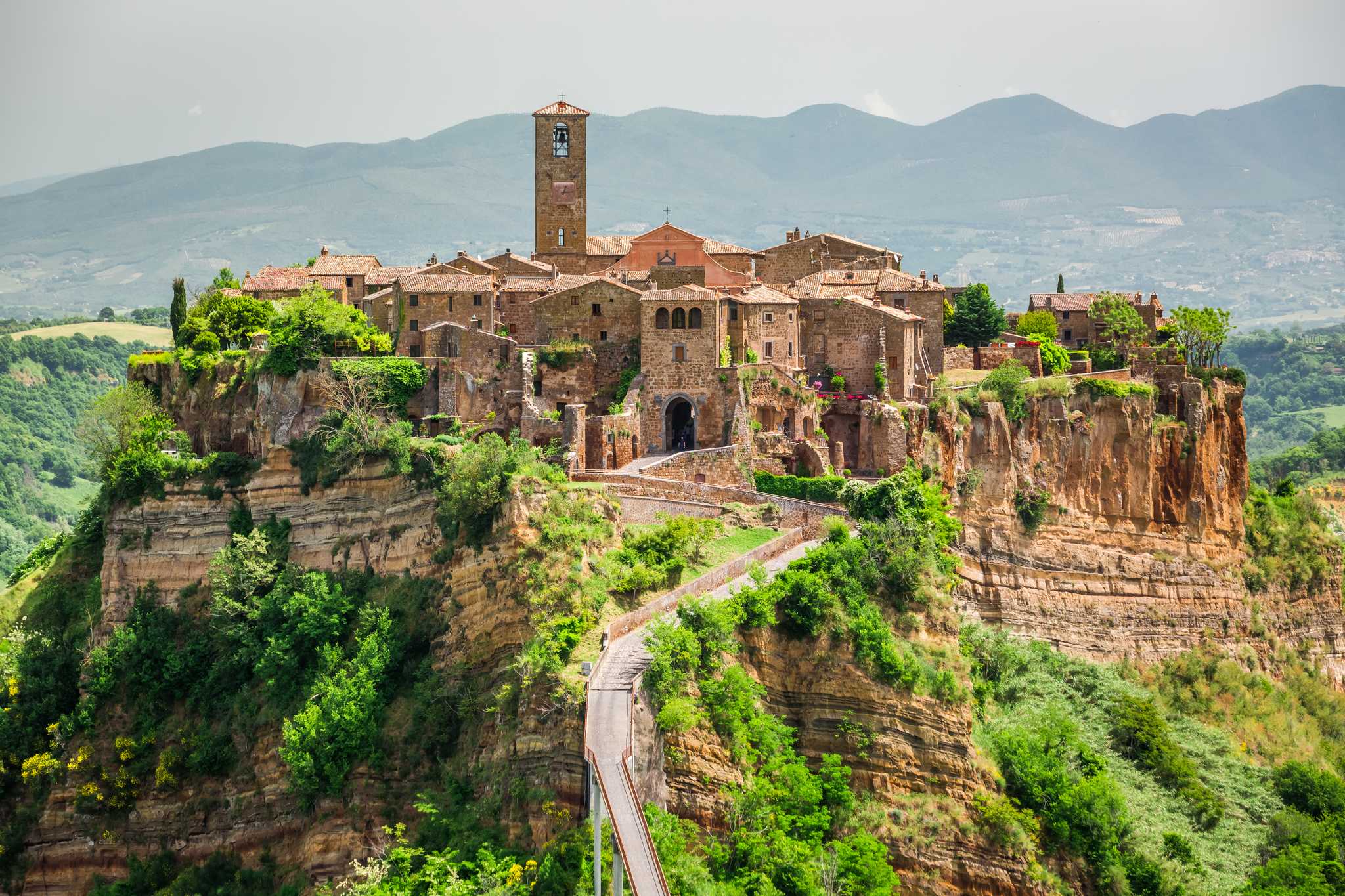Civita di Bagnoregio, a stone hilltop town on eroded cliffs with a footbridge and mountains behind.