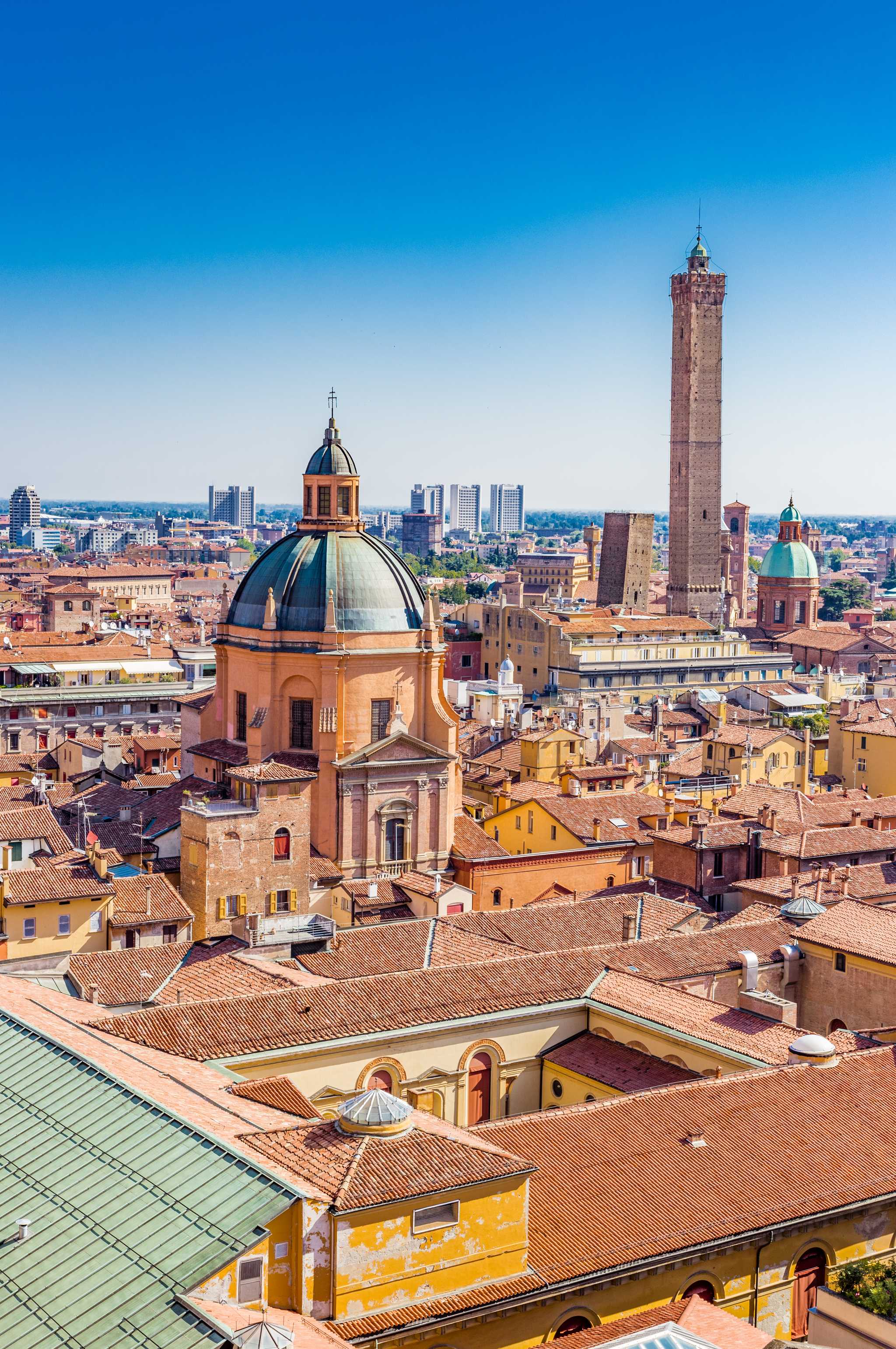 A rooftop view over Bologna, a domed church and the leaning medieval Asinelli tower above terracotta roofs.