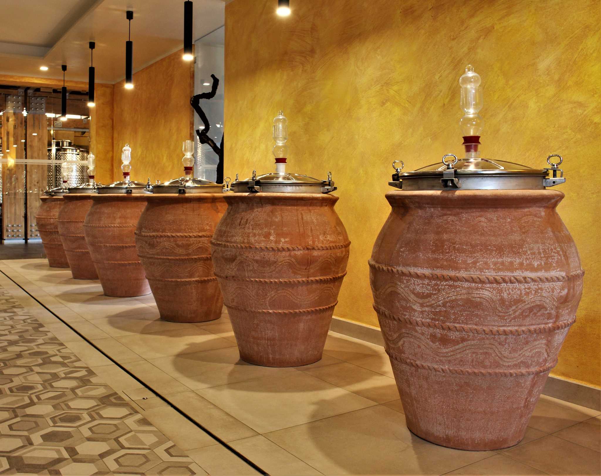 Row of large terracotta wine amphorae with metal lids at Bosco de' Medici, against a gold wall.