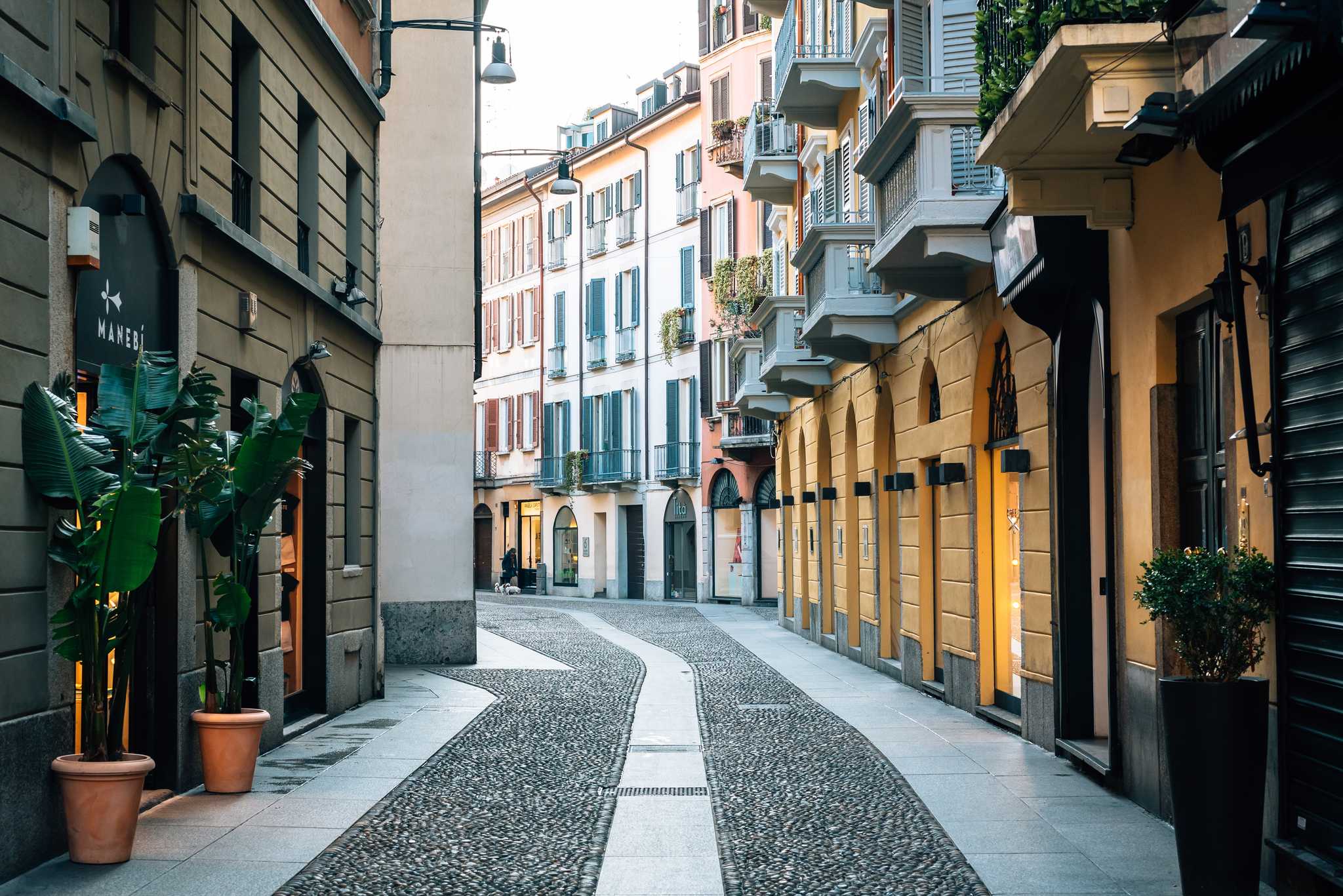 A cobblestone street of colorful storefronts with plants in planters.
