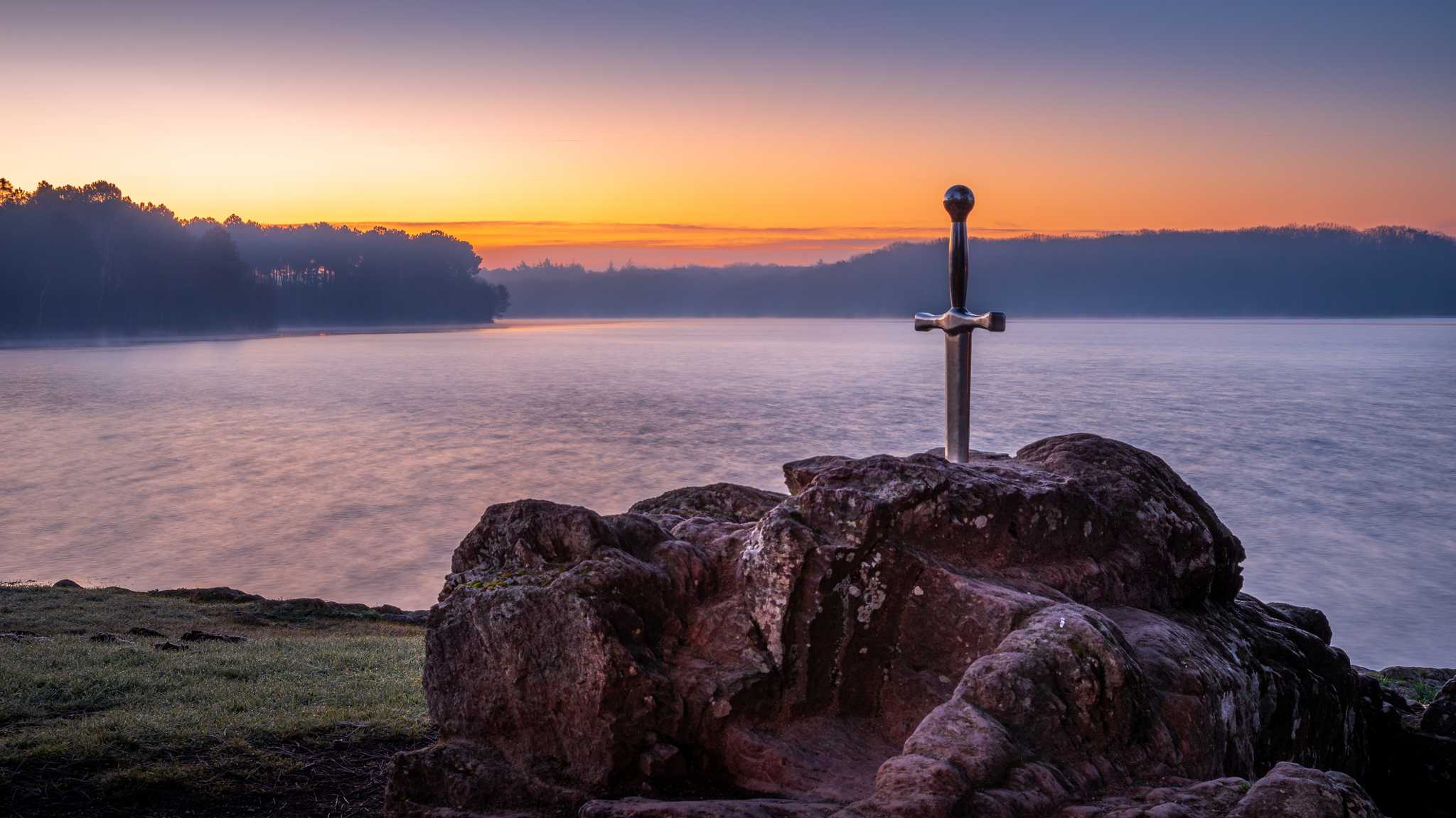 A silver sword embedded in a boulder on the water’s edge at sunset, with forests beyond.