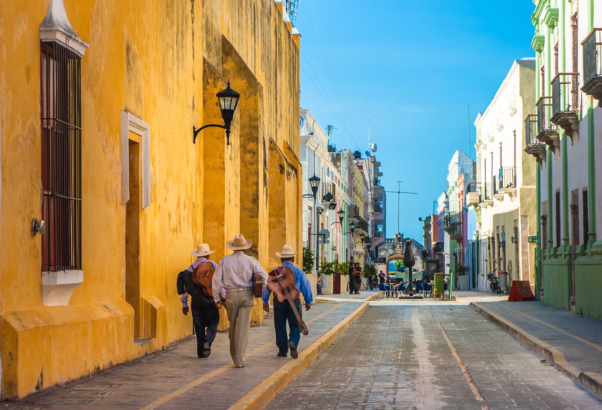 Three men with guitars walk down a street of brightly colored buildings in Campeche.