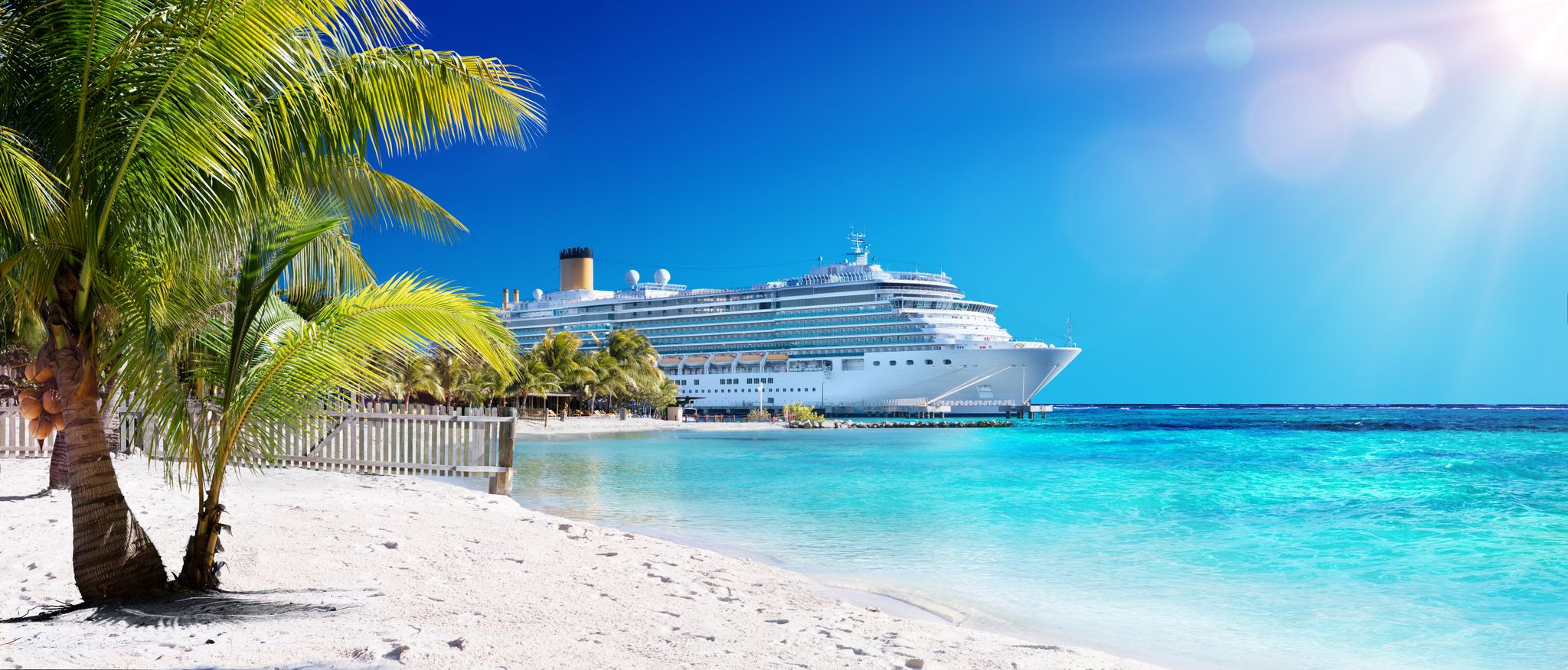Cruise ship docked beside a white sand beach with palm trees and turquoise water.