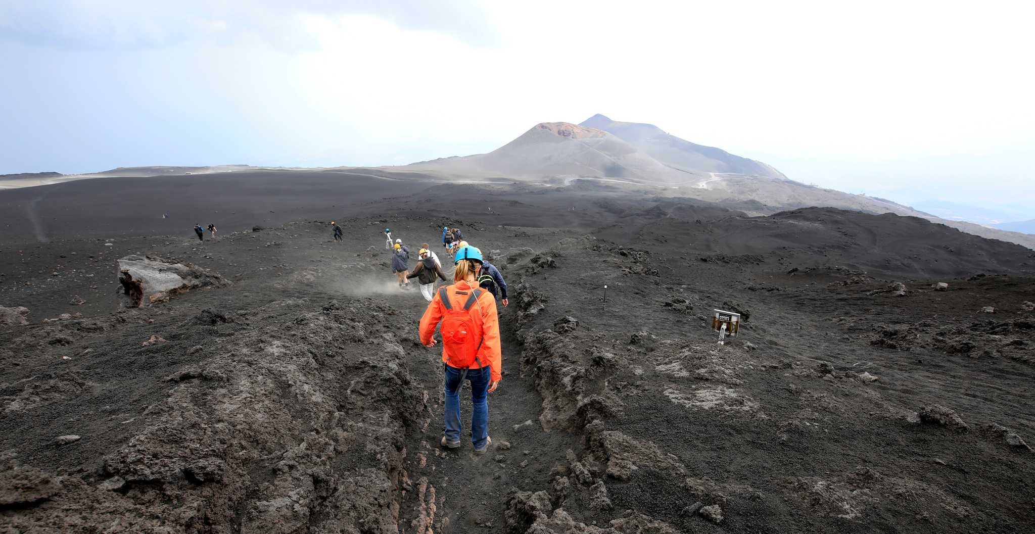 Hikers climbing a black volcanic trail toward Mount Etna's summit craters, Sicily.