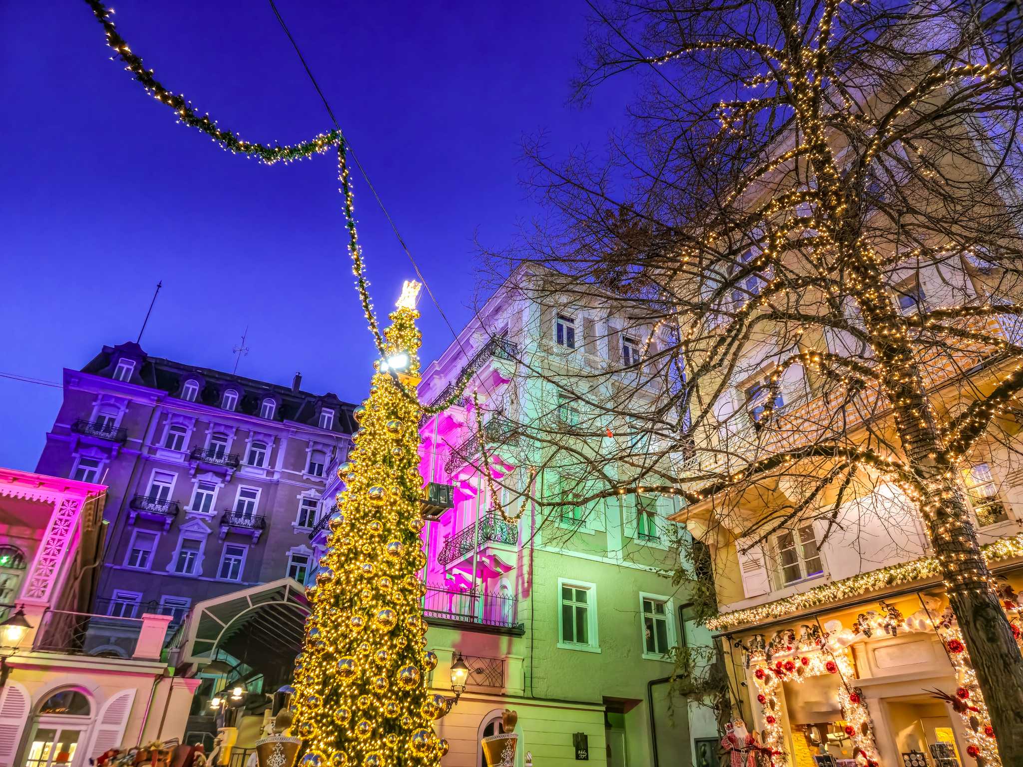 A lighted Christmas tree in front of buildings lit by pink, purple, and green spotlights.