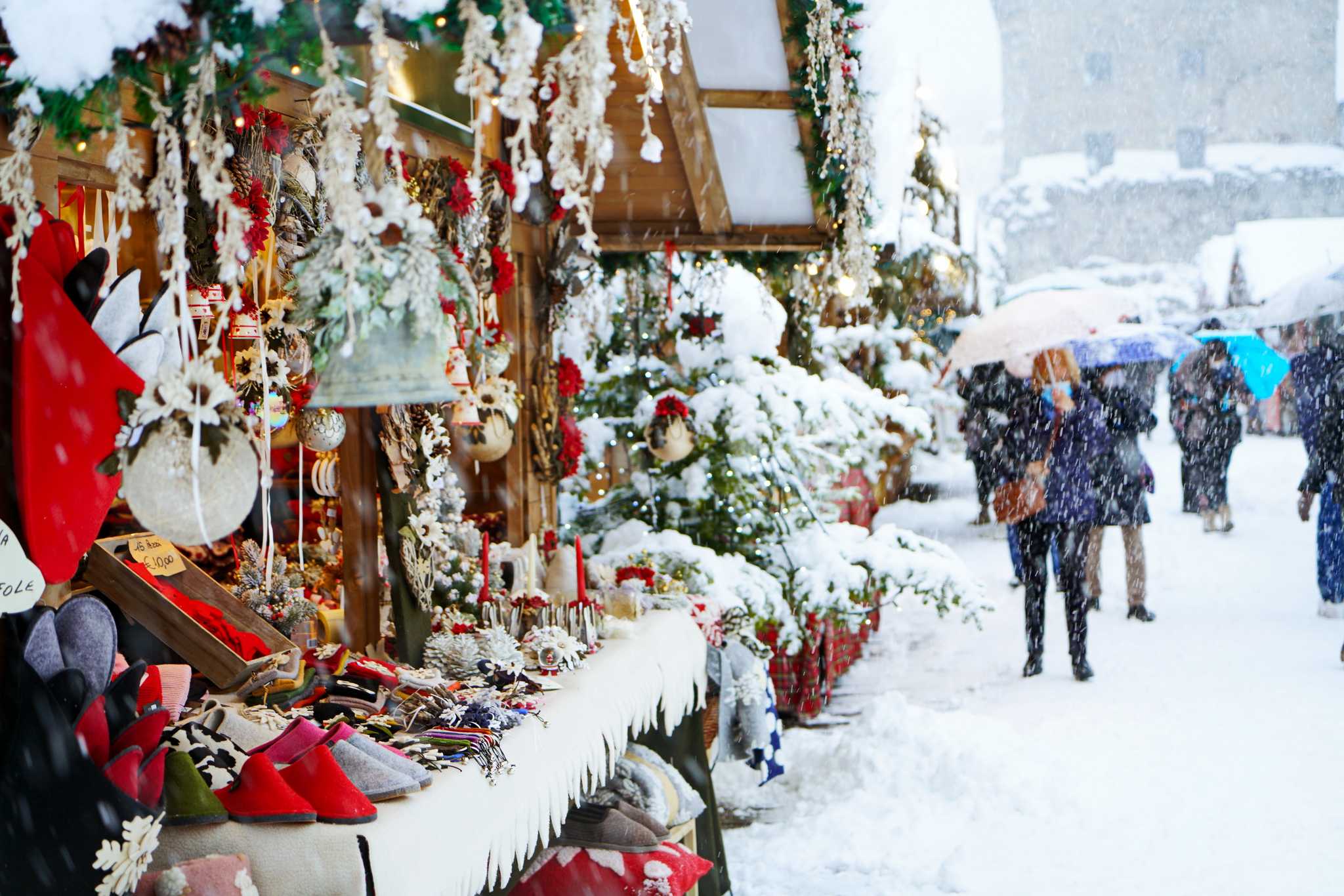 Heavy snowfall at a Christmas market stall in Aosta, people walking past on the right.