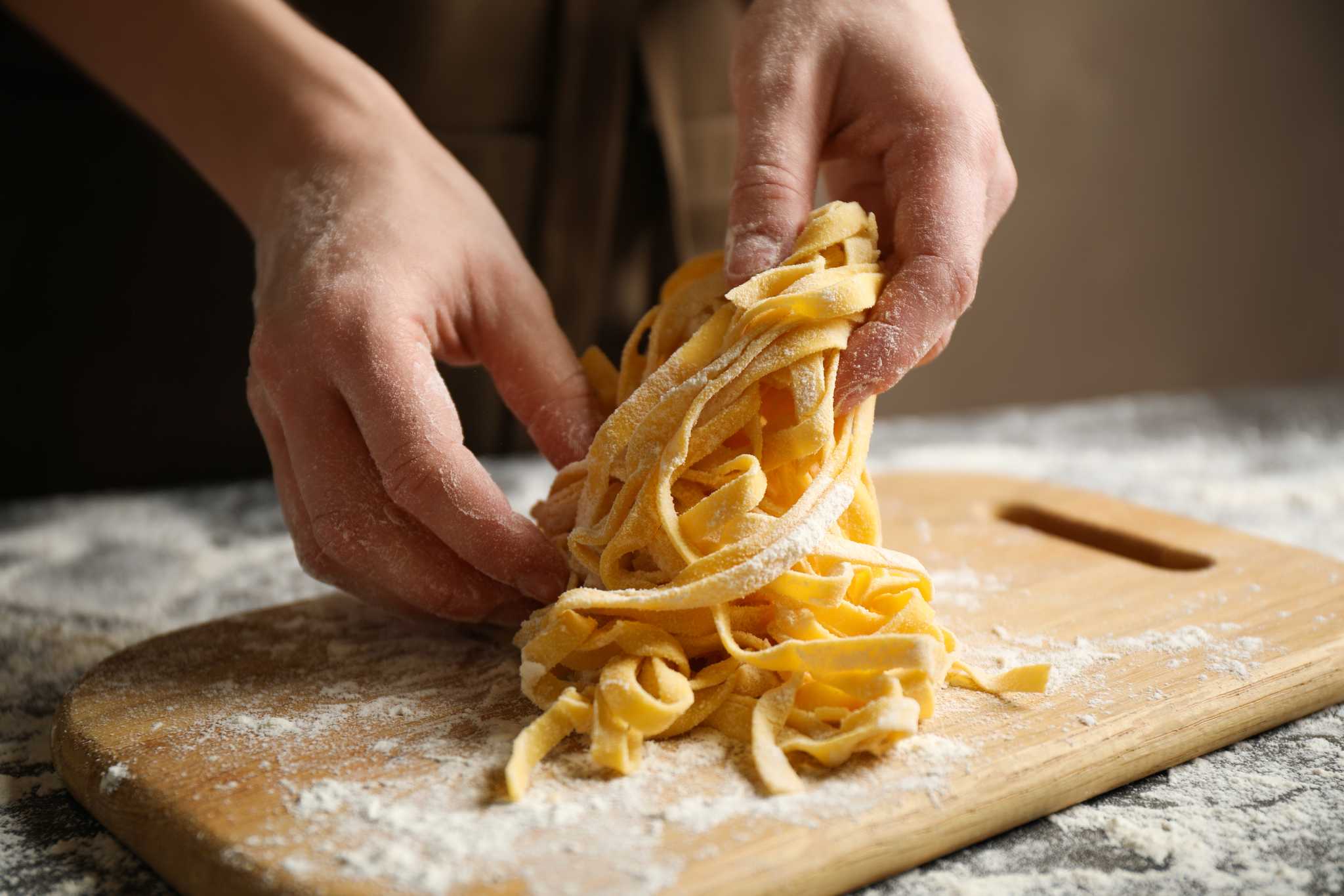 Hands lifting fresh-cut ribbons of homemade egg pasta from a flour-dusted cutting board.