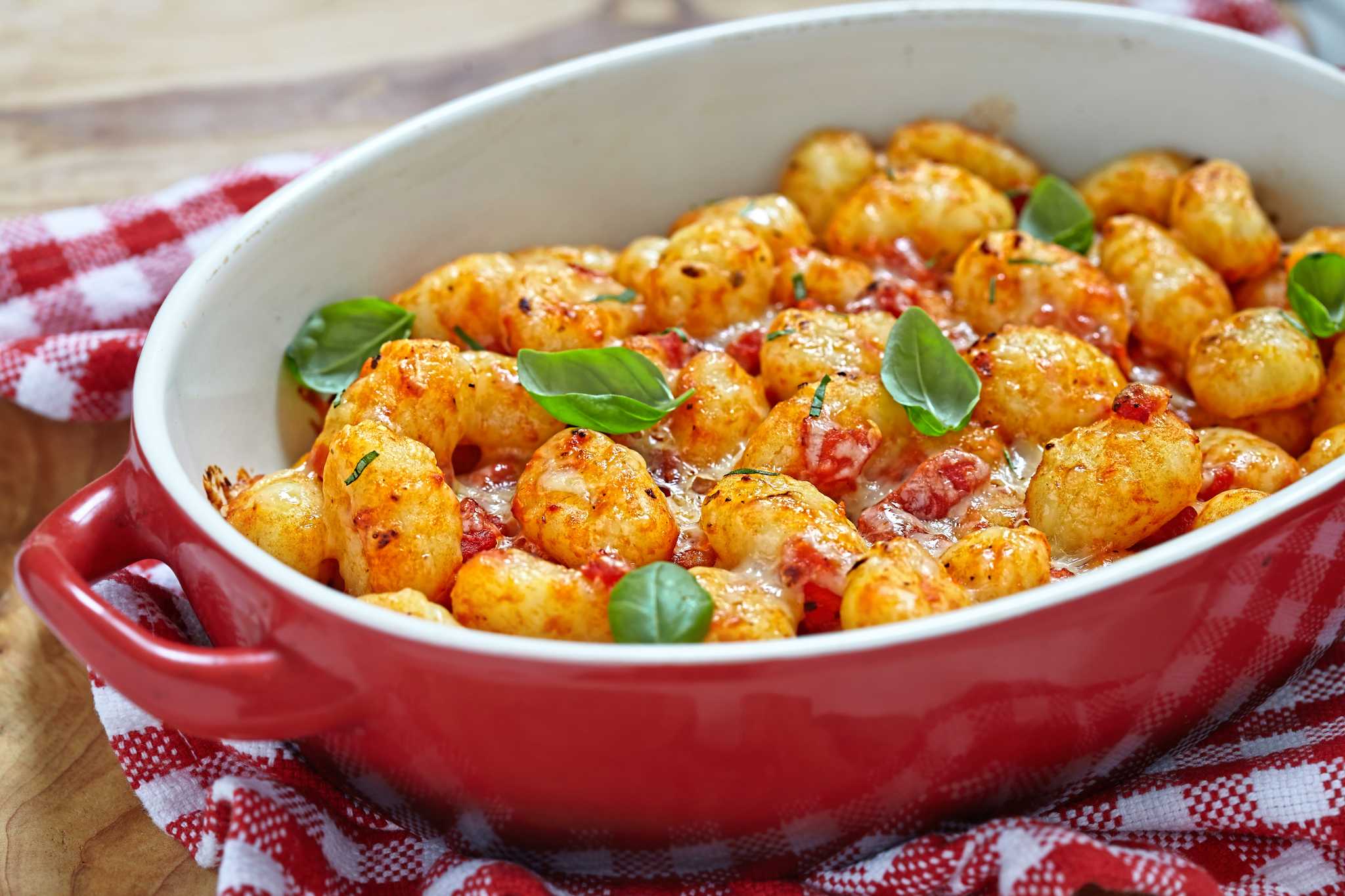 A red-and-white oval baking dish of golden baked gnocchi in tomato sauce.