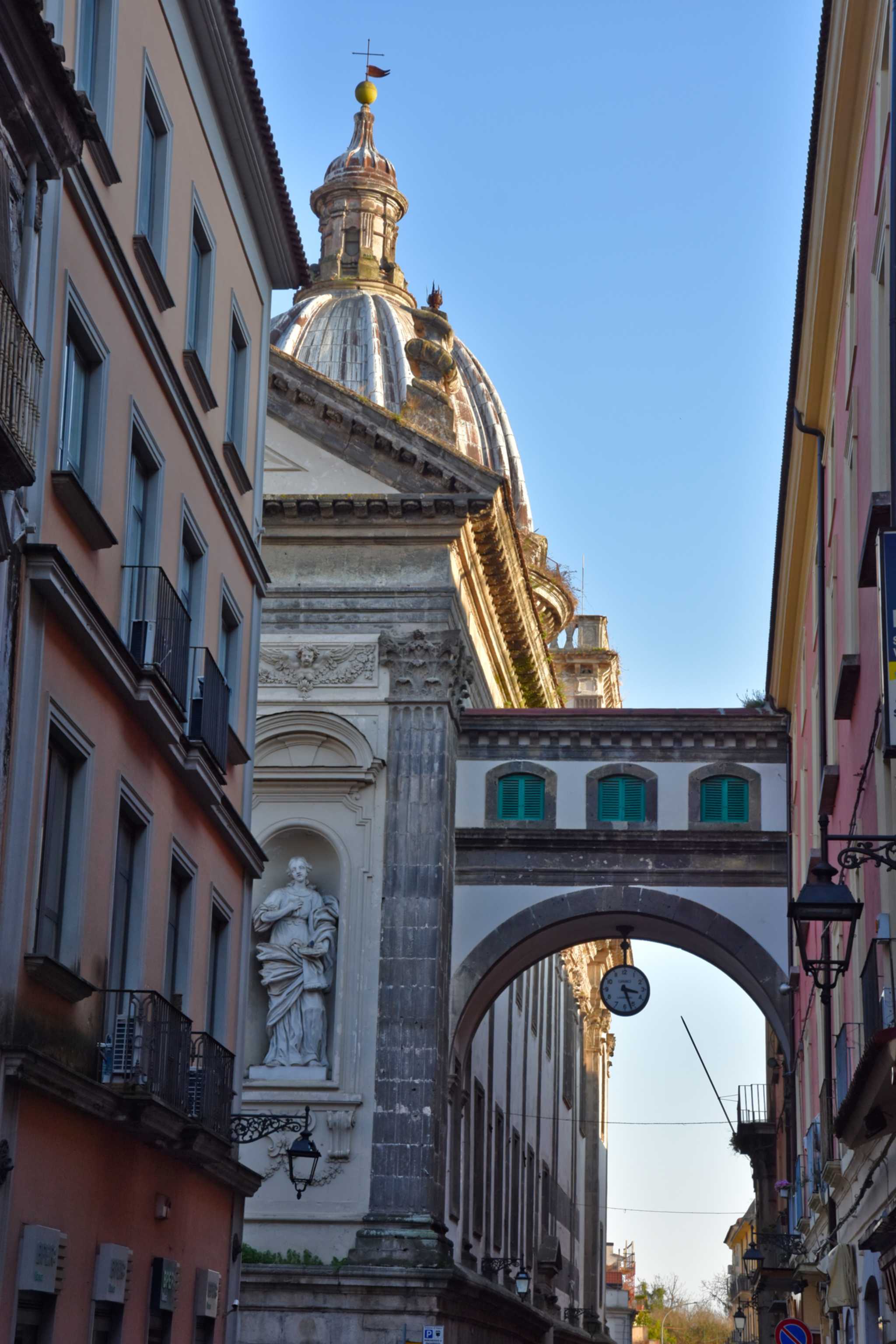 An arch with a clock over an old town street in Capua, a carved statue on its left and a domed building behind.