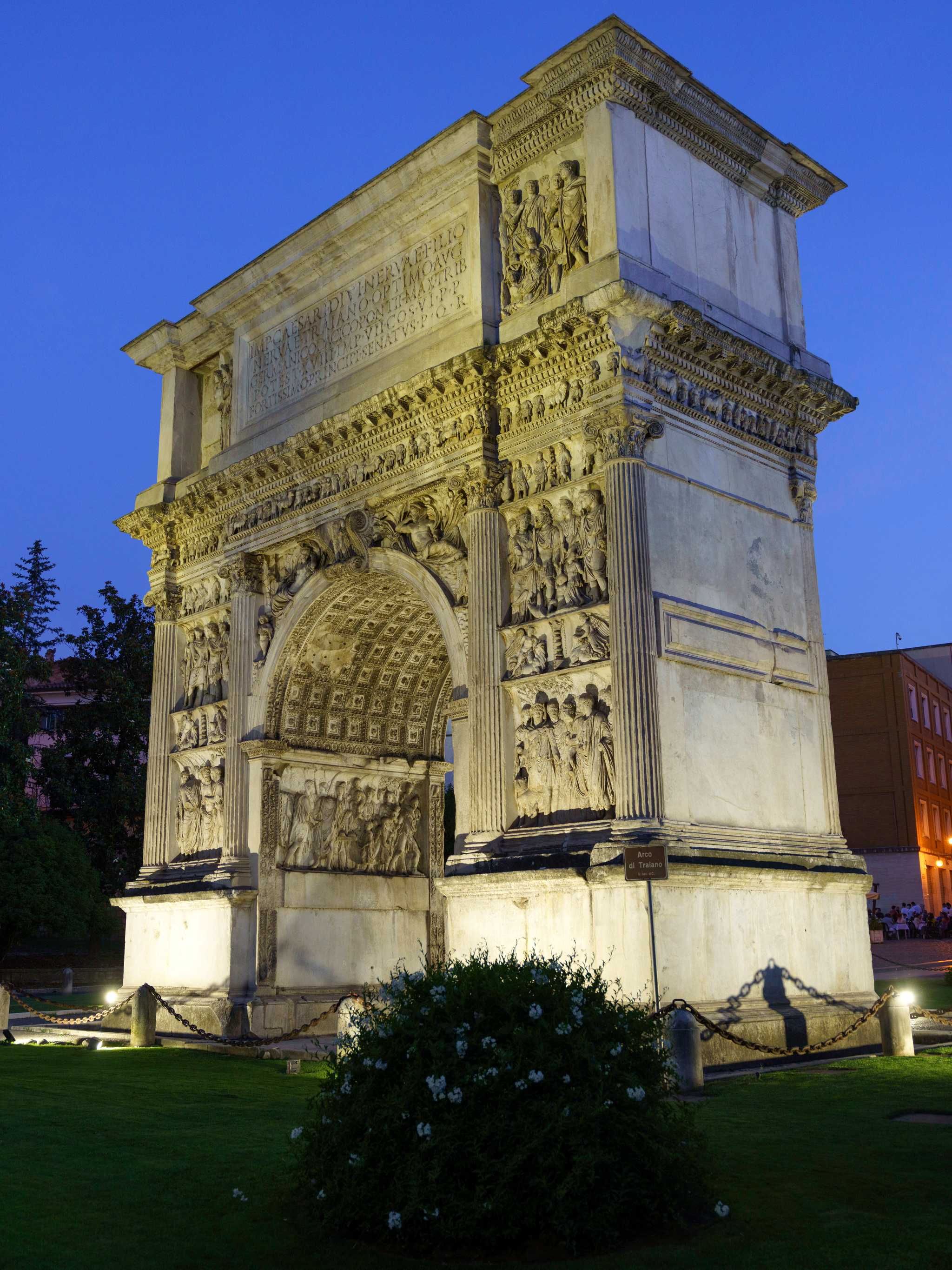 The Roman Arco di Traiano at night, lit up, with grass and a bush in the foreground.