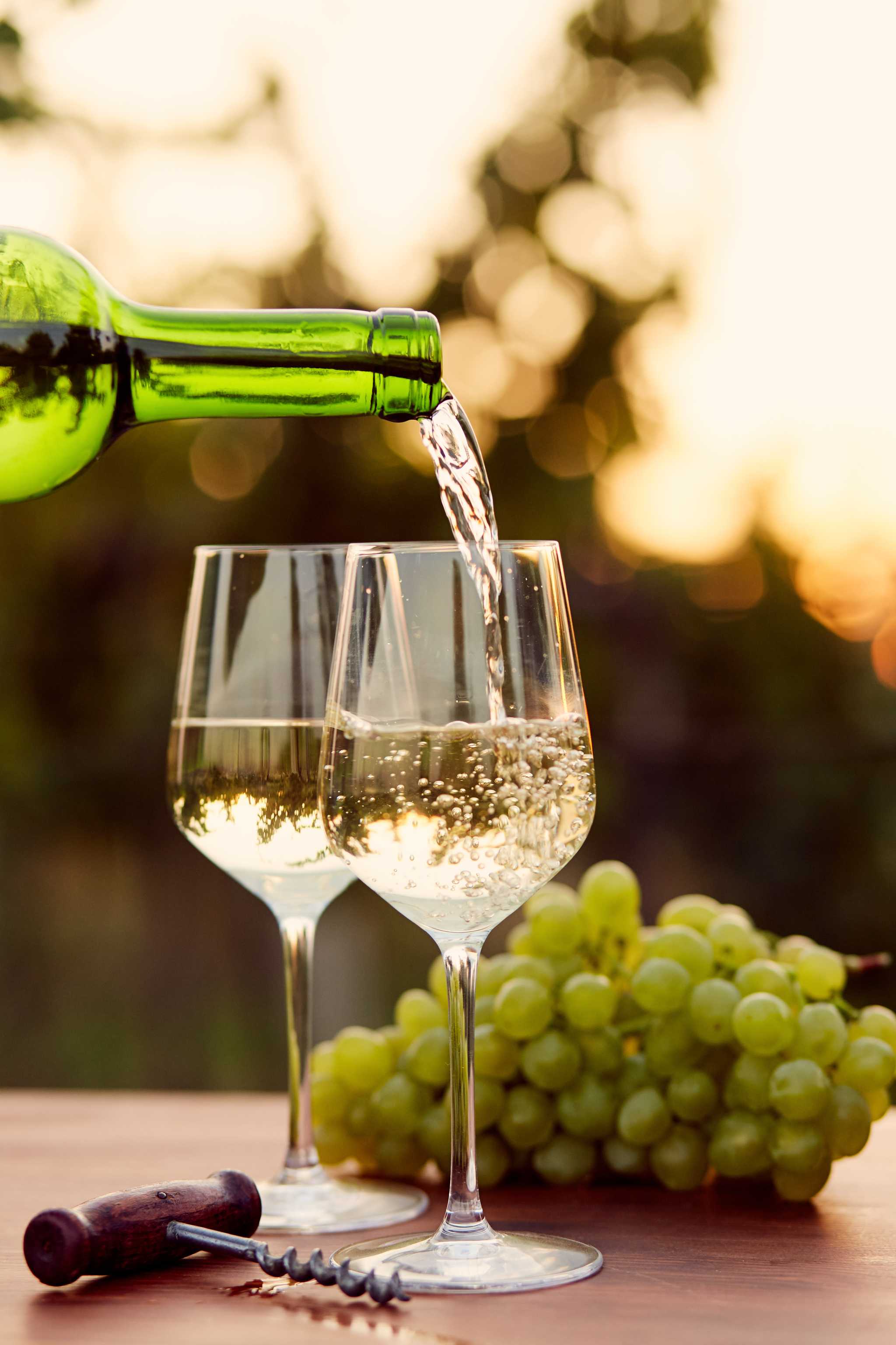 Two glasses of white wine on a wooden table with green grapes, one glass being filled by a green bottle.