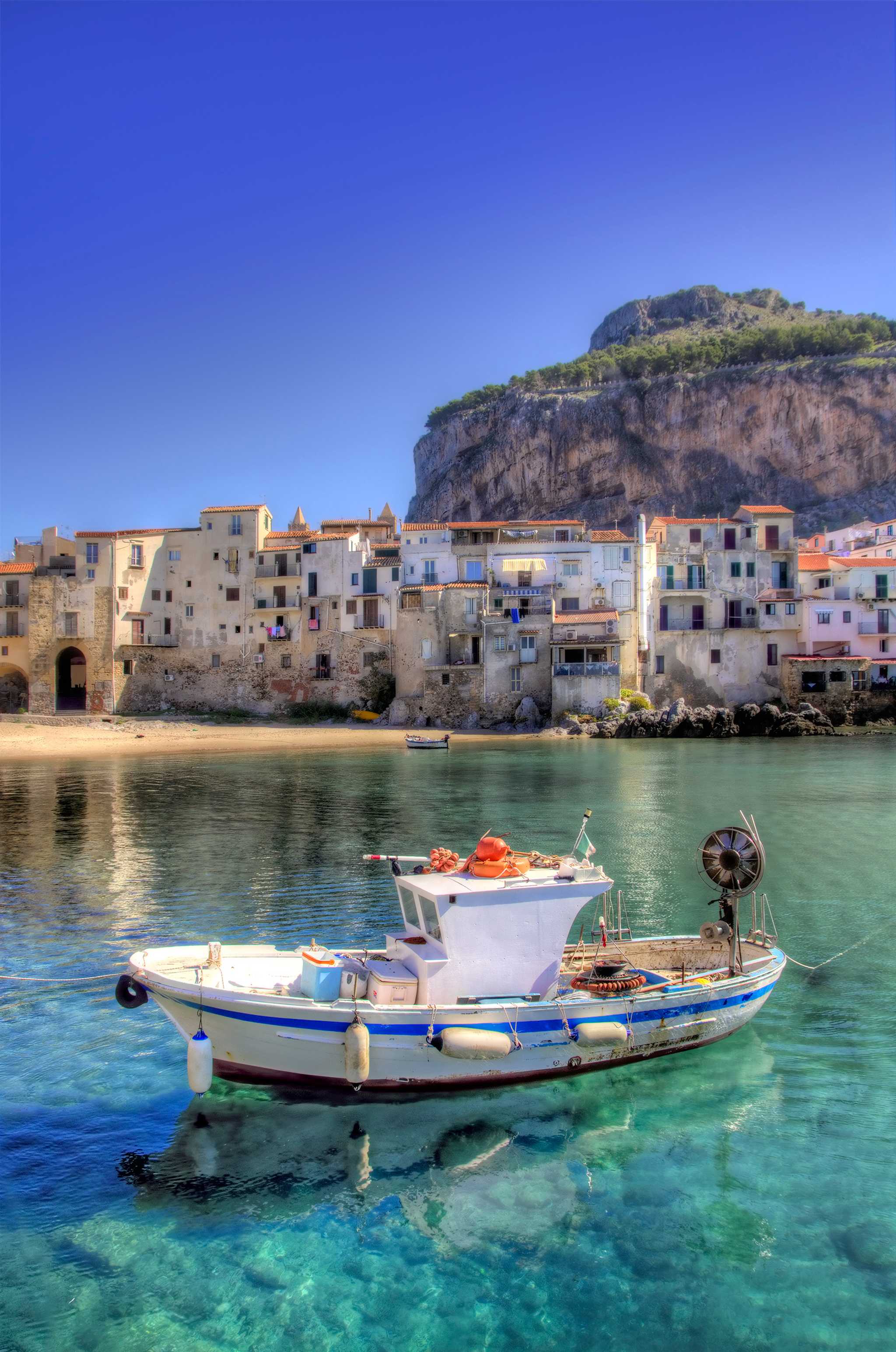 Fishing boat floating in clear blue water in CefalĂš, with traditional homes behind.