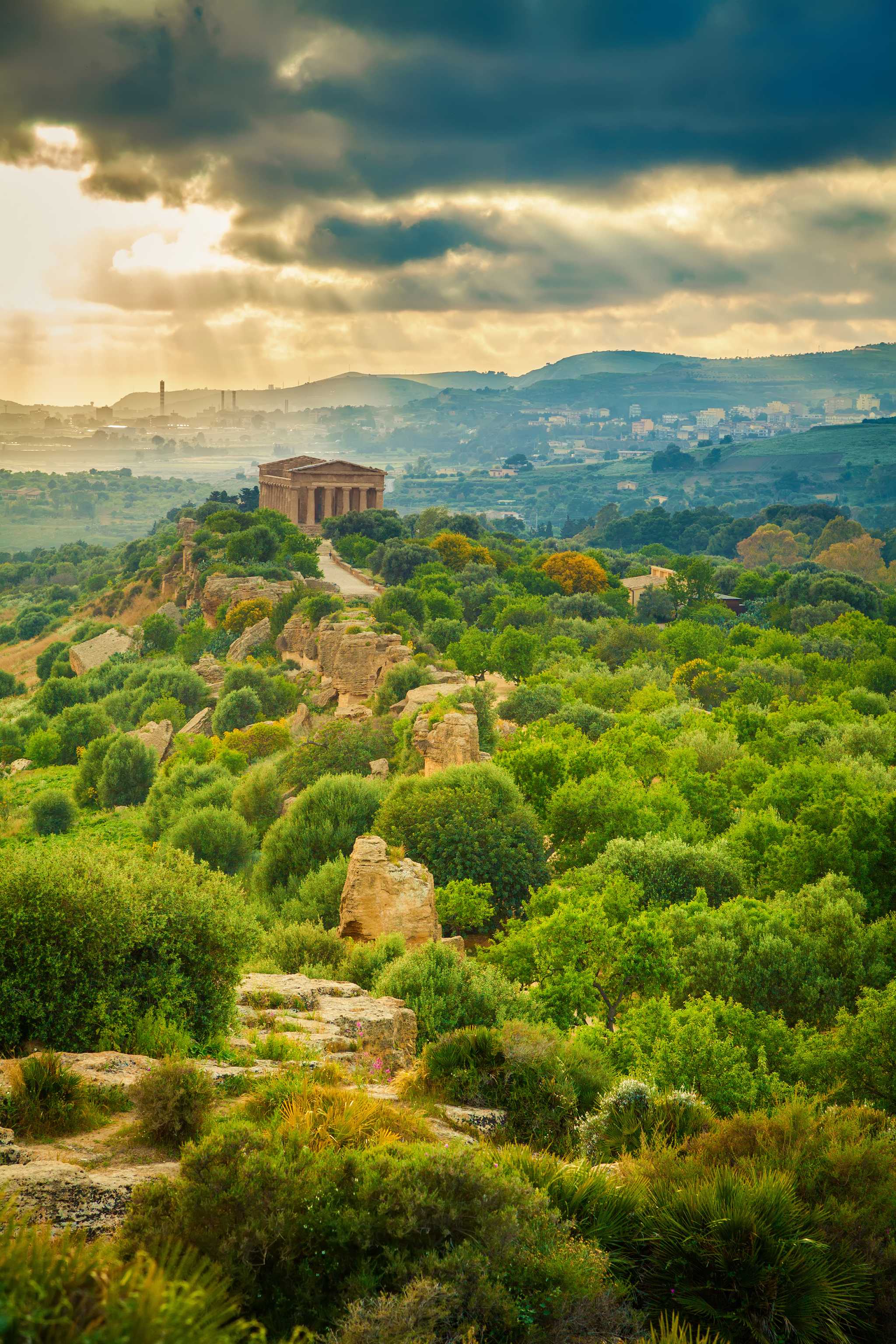 Ancient Greek temple on a hill surrounded by lush greenery, sunlight breaking through dark clouds.