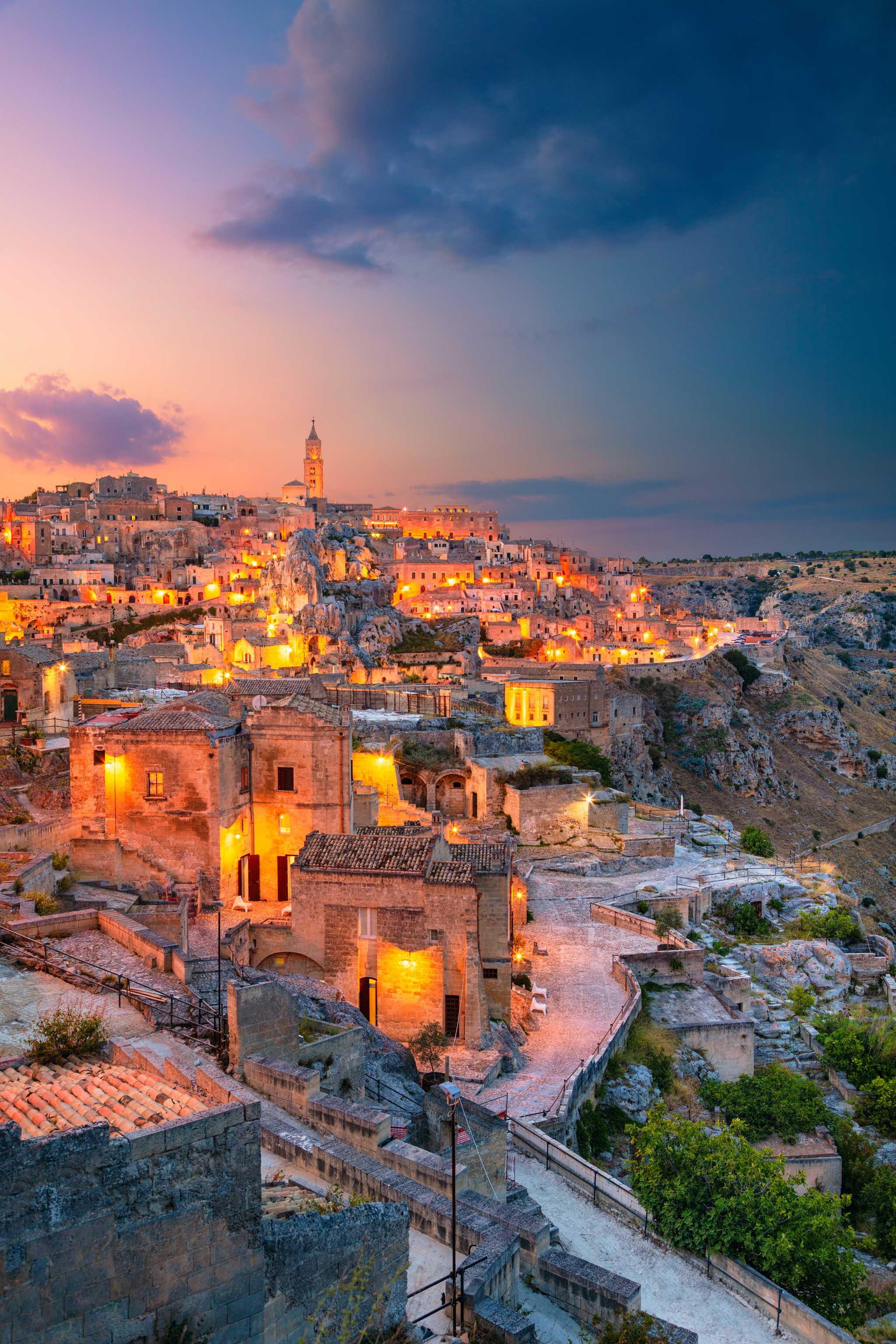 Matera during a cloudy sunset with bright orange lights on in many buildings.
