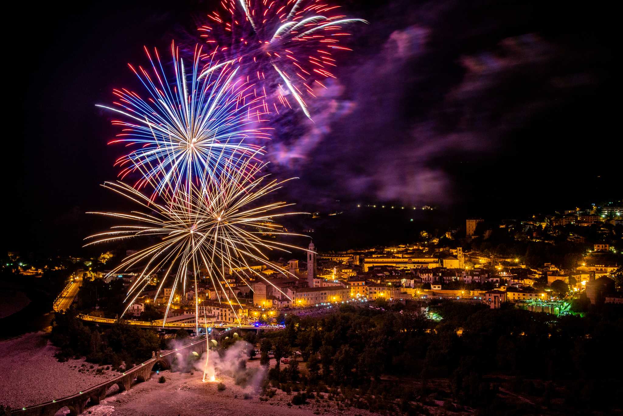 Ferragosto fireworks burst over the lit town of Bobbio and its bridge at night.
