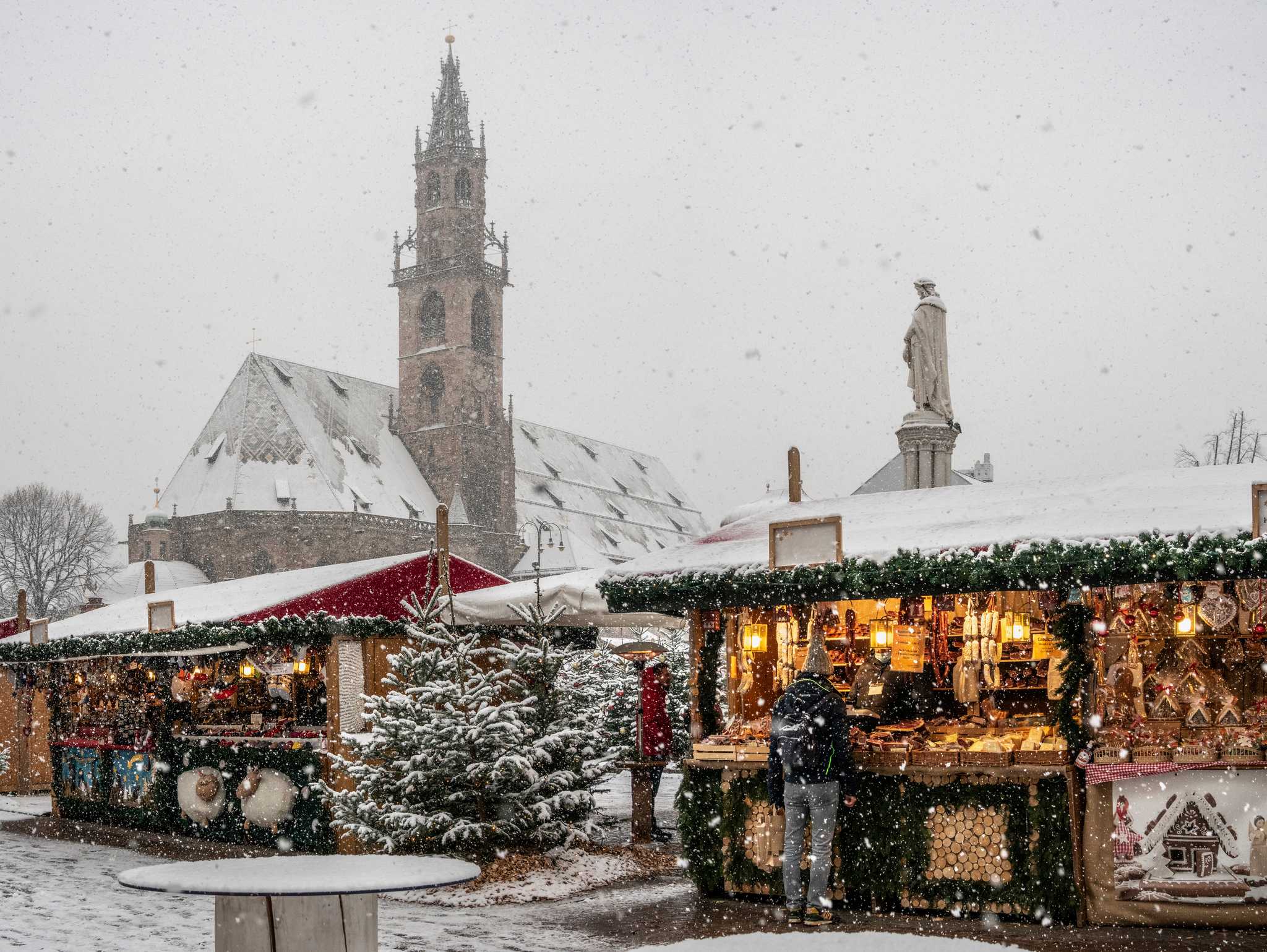 Snowy Christmas market stalls before Bolzano's cathedral spire, shoppers browsing lit huts.