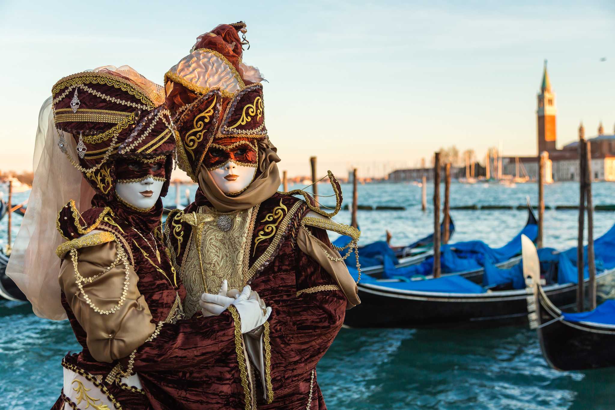 Two people in ornate Venice Carnival masks and costumes pose by gondolas on the lagoon.
