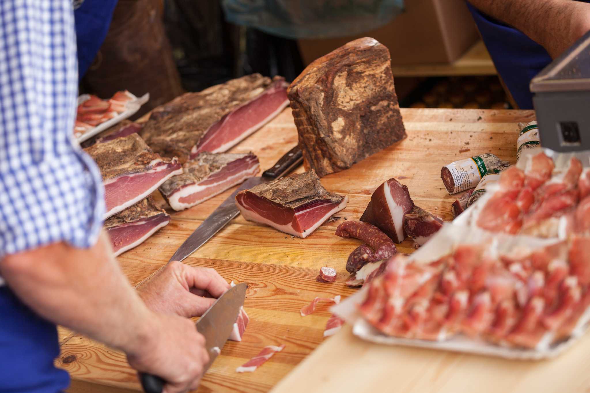 Man in blue checkered shirt thinly slicing Speck on a wooden butcher block.