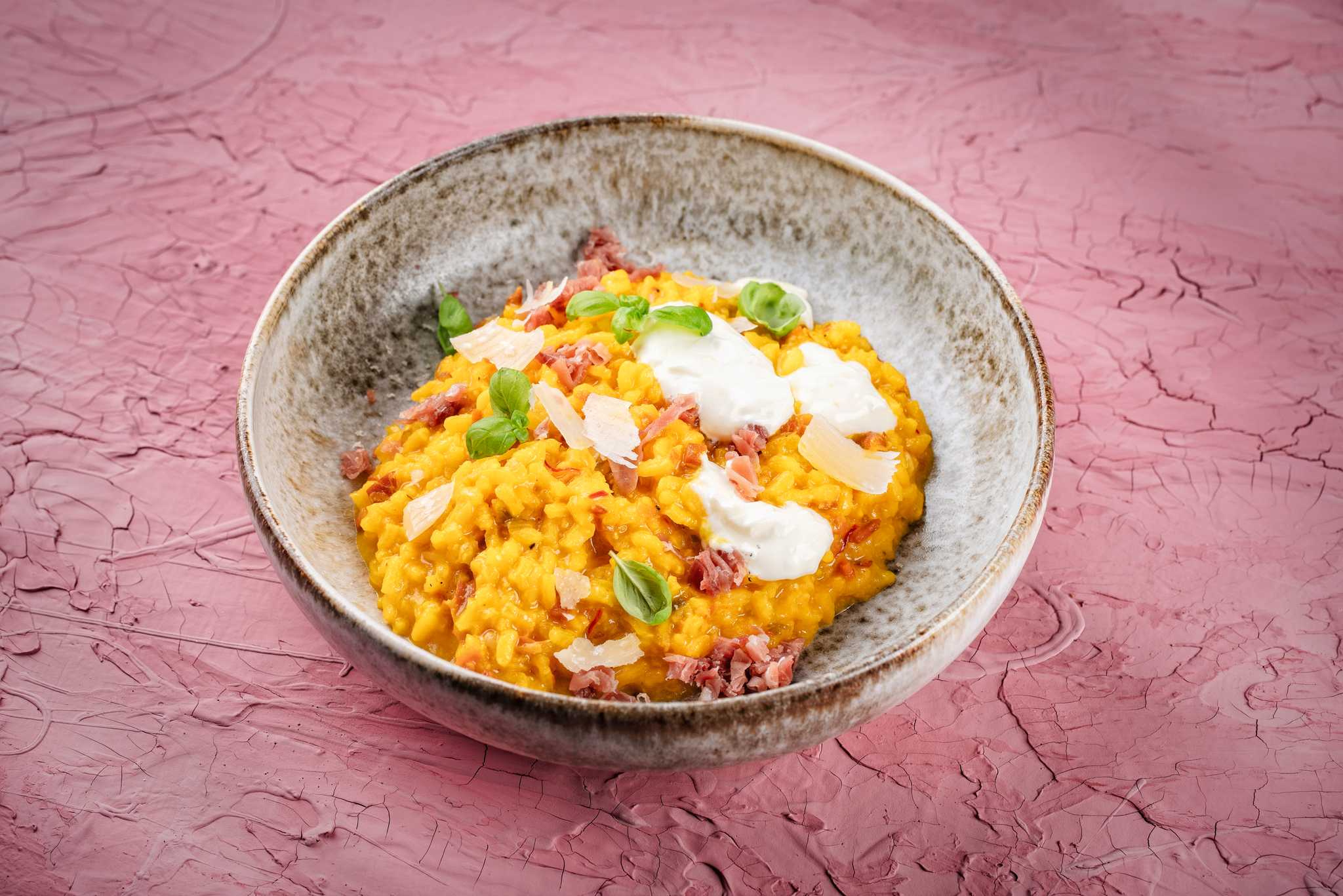 Bowl of yellow risotto alla Milanese topped with prosciutto, herbs, and cheese on a pink background.