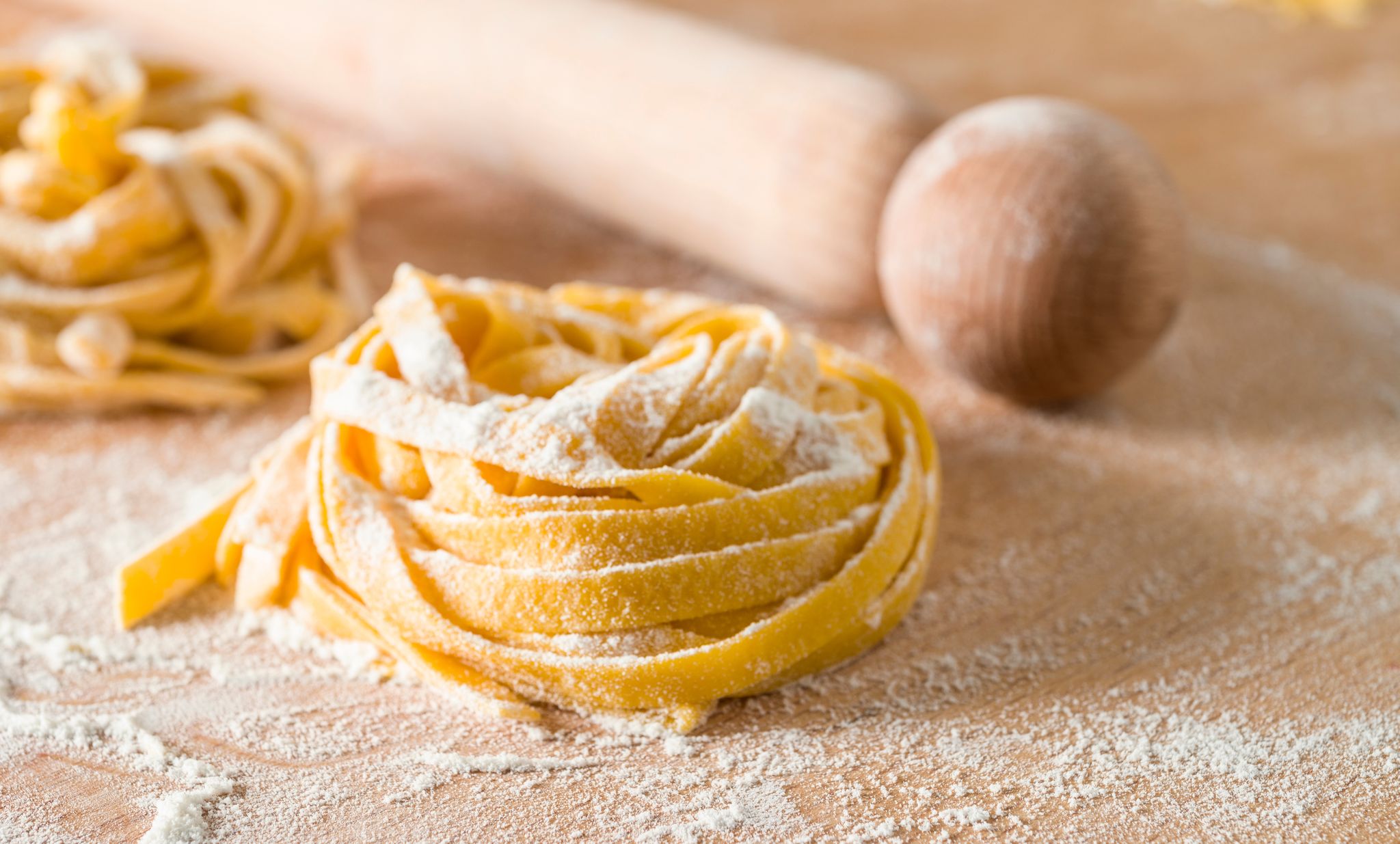 Two small piles of fresh linguine noodles coated in flour beside a wooden rolling pin.