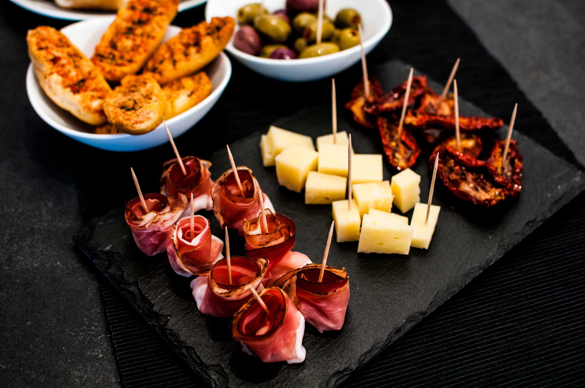 Buffet of aperitivo snacks, including cured meats, cheeses, and olives, on a black table.
