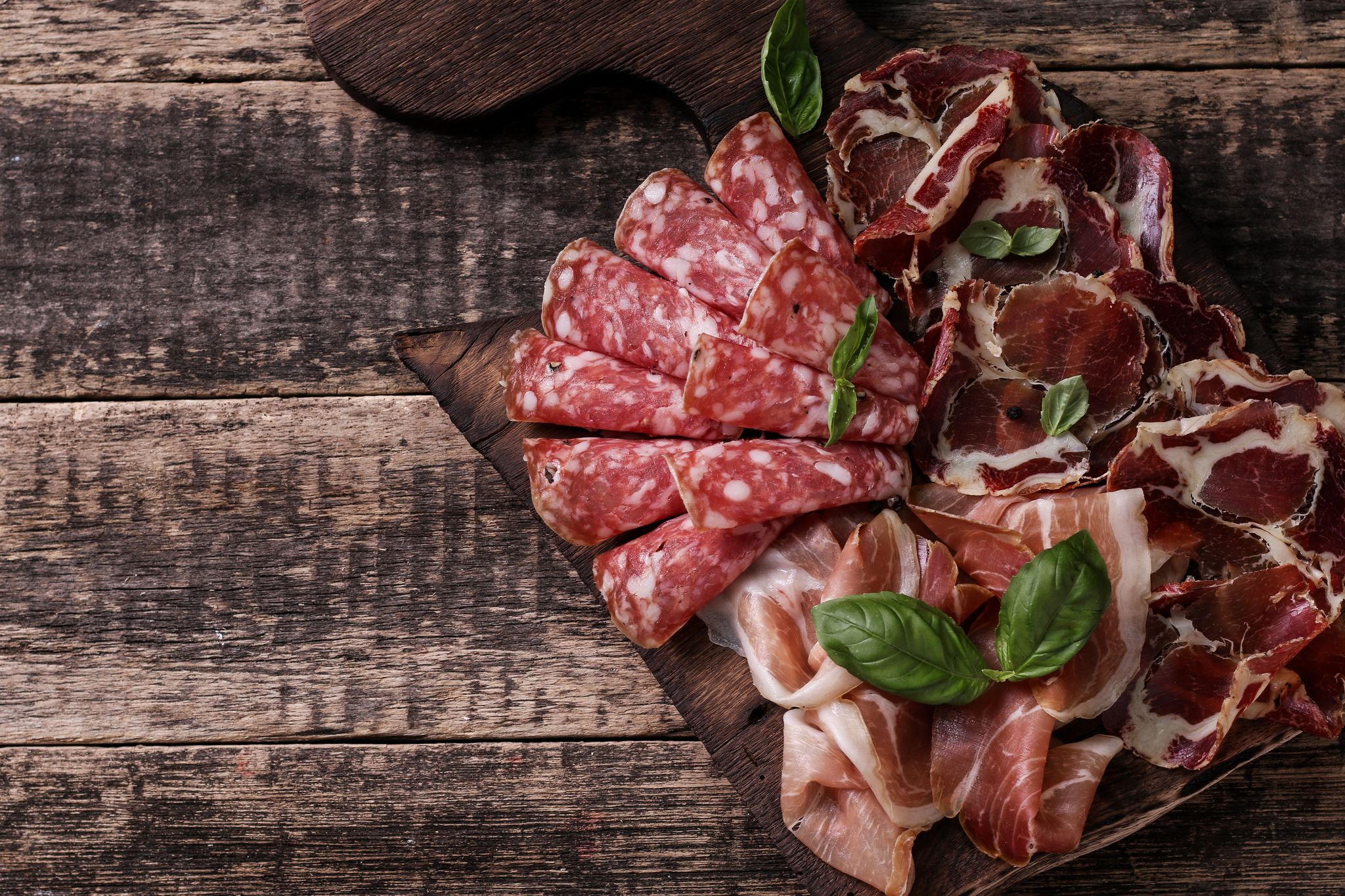 Variety of cured meats on a wooden board with basil garnish.